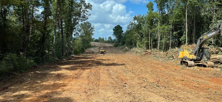 Heavy machinery clears a wide dirt path through a forest under a blue sky with clouds.