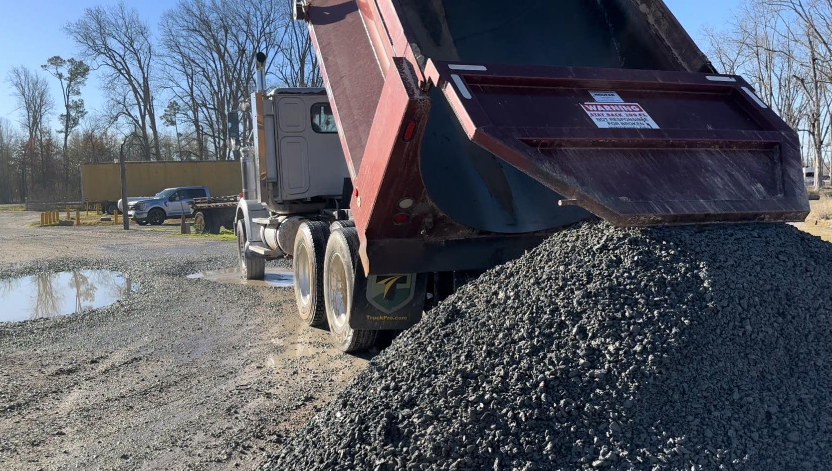 A red dump truck tilted back, unloading a large pile of gravel in an outdoor yard on a sunny day.