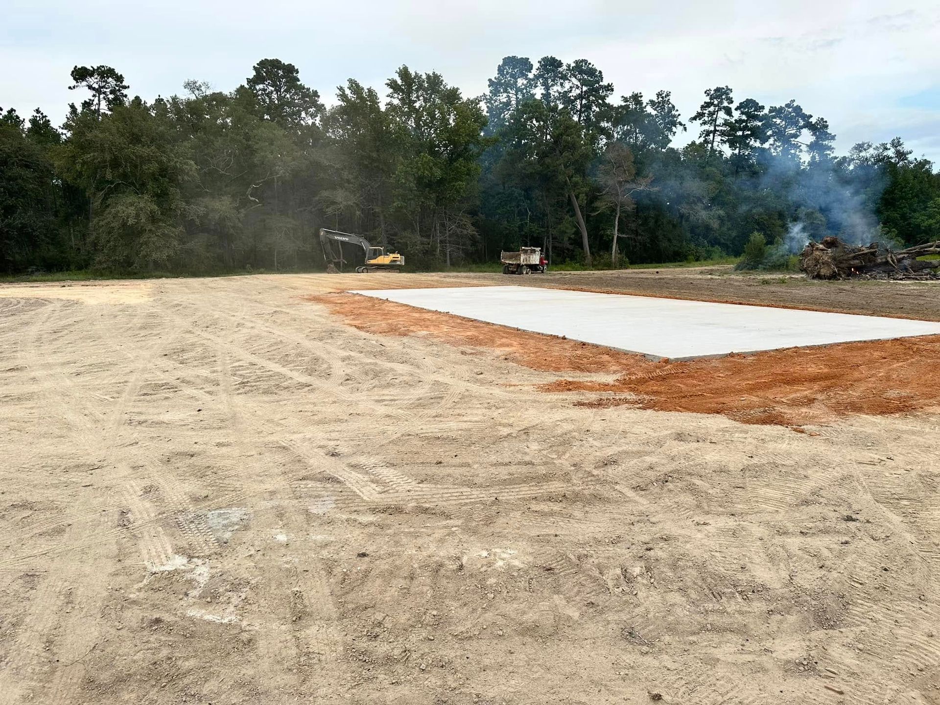 A concrete slab foundation sits on a cleared, sandy construction site with heavy machinery working near a treeline.