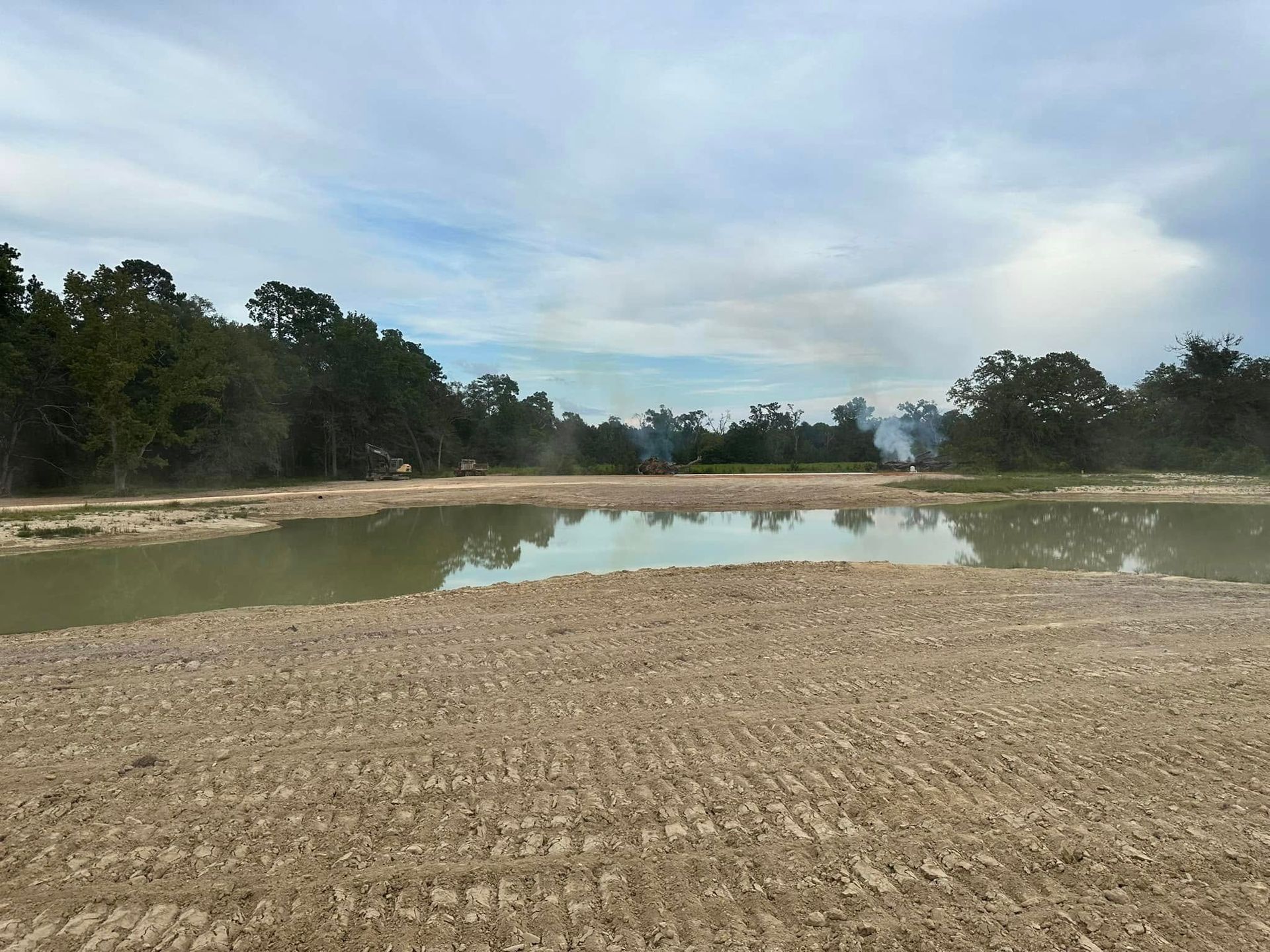 A muddy, cleared construction site with a large, calm pool of water reflecting the cloudy, light blue sky and distant trees.