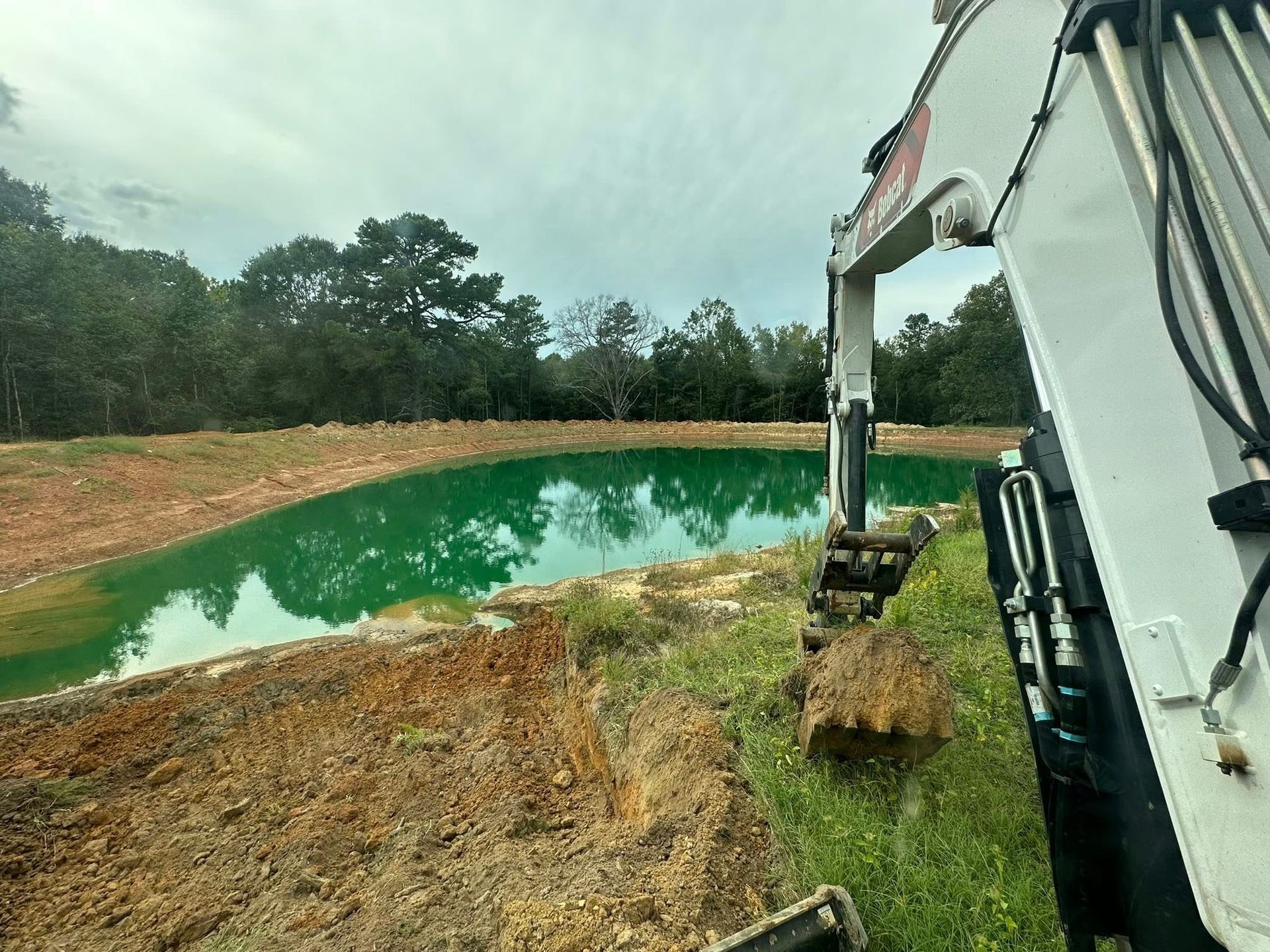 An excavator arm sits near a small, bright green pond bordered by dirt and trees under a cloudy sky.