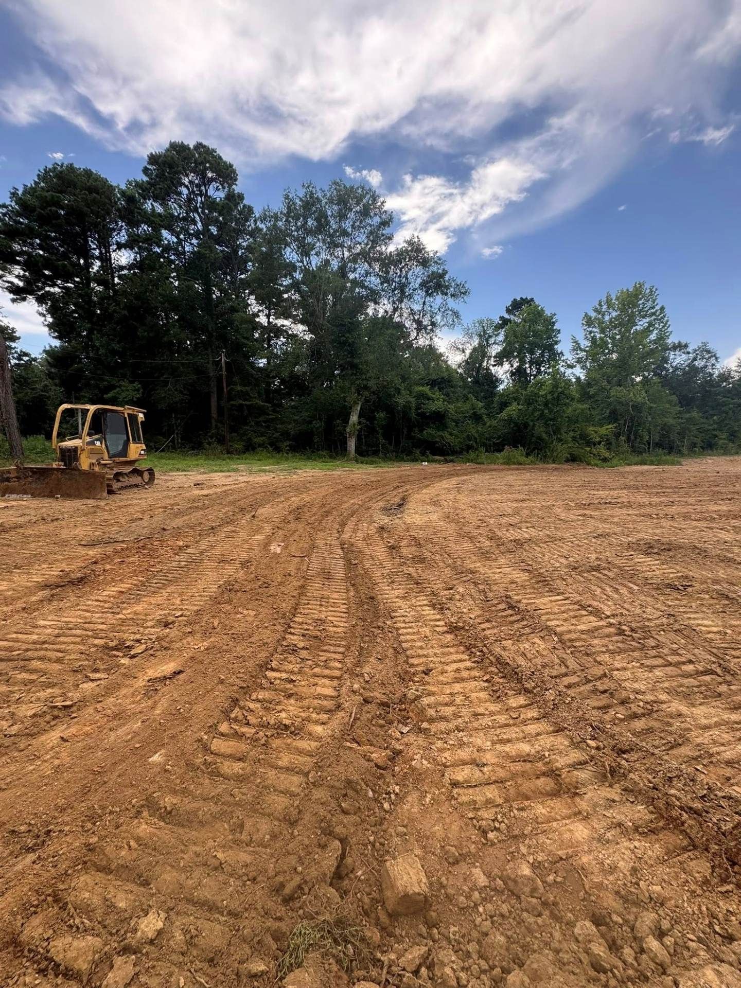 A yellow bulldozer sits on a clearing of churned brown earth next to a line of trees under a bright blue sky.