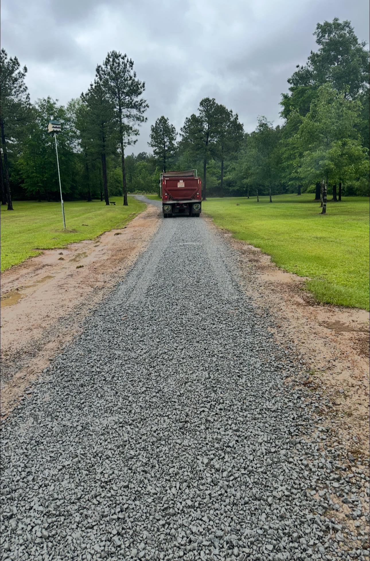 A red dump truck spreading gray gravel on a dirt driveway lined by green trees and grass under a cloudy sky.
