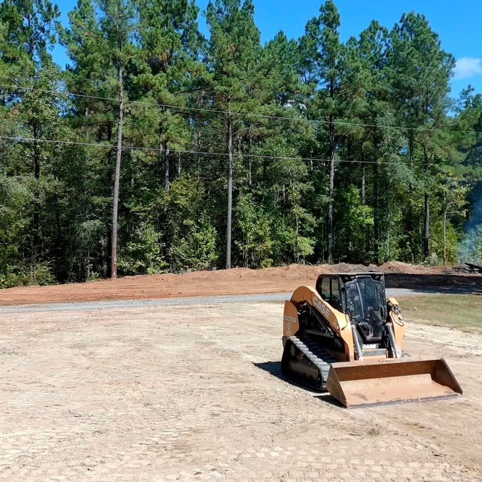 A yellow skid-steer loader sits on a leveled dirt lot in front of a dense treeline on a clear, sunny day.