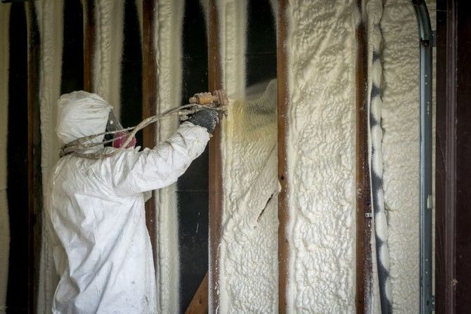 Worker in white protective suit spraying insulation foam inside wall studs