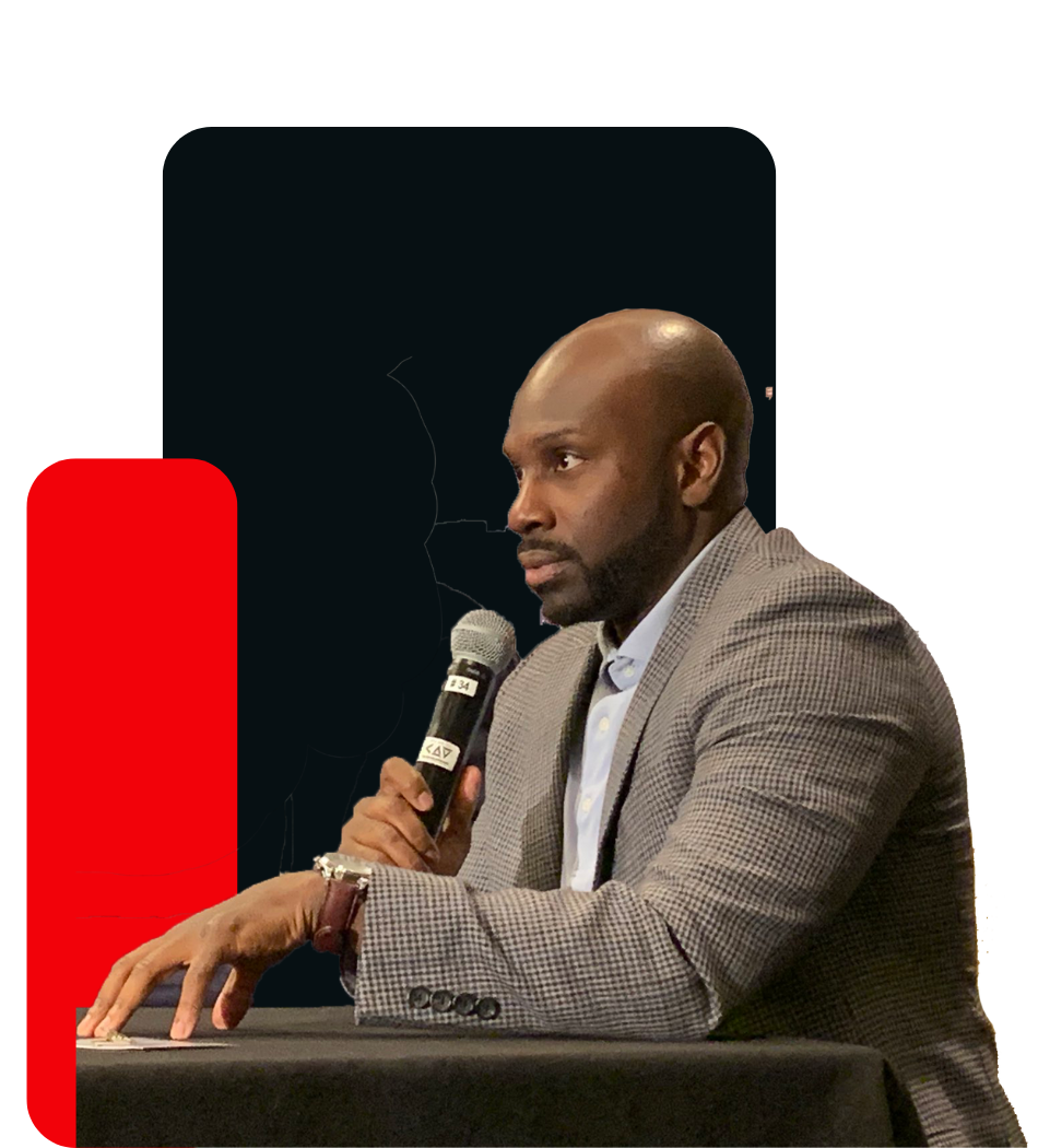 A Juntae Delane in a suit is sitting at a table holding a microphone.