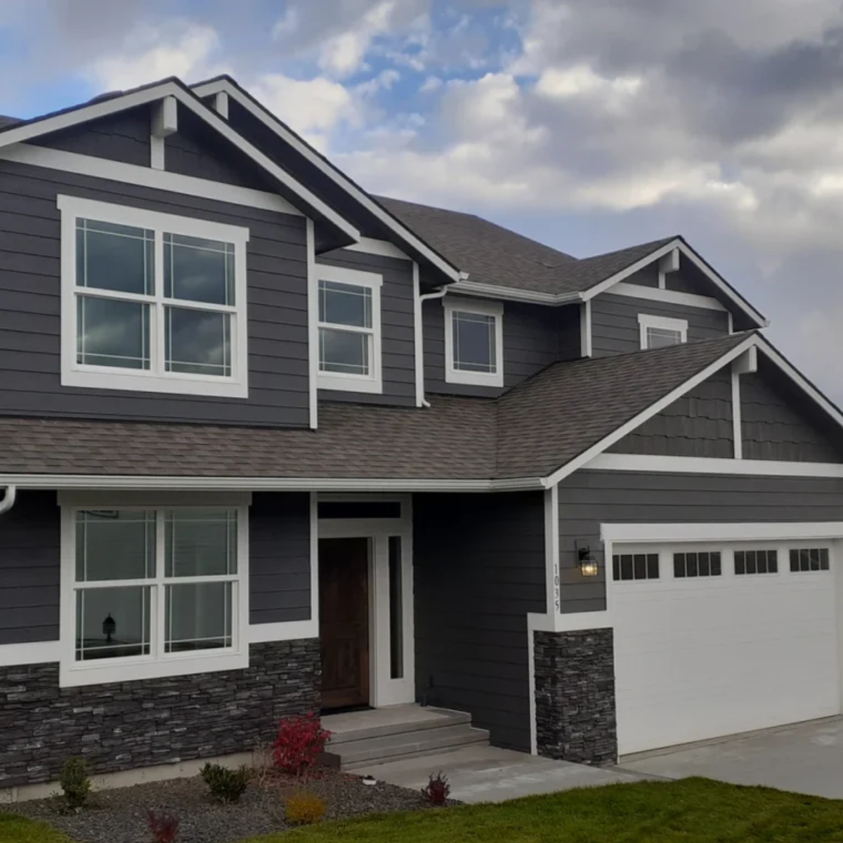 Two-story gray house with white trim, stone accents, and a white garage door under a cloudy sky.