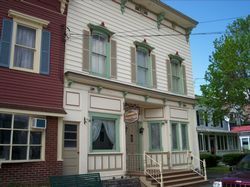 Two-story cream-colored building with green trim, three windows on the second floor, a small porch, and a red building to the left.