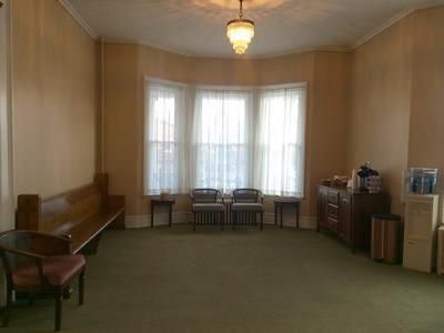 Empty room with a bay window, wood bench, chairs, cabinet, and water cooler; neutral color scheme.