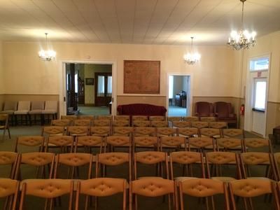 Rows of chairs face a small stage, in a room with chandeliers and exit doors.