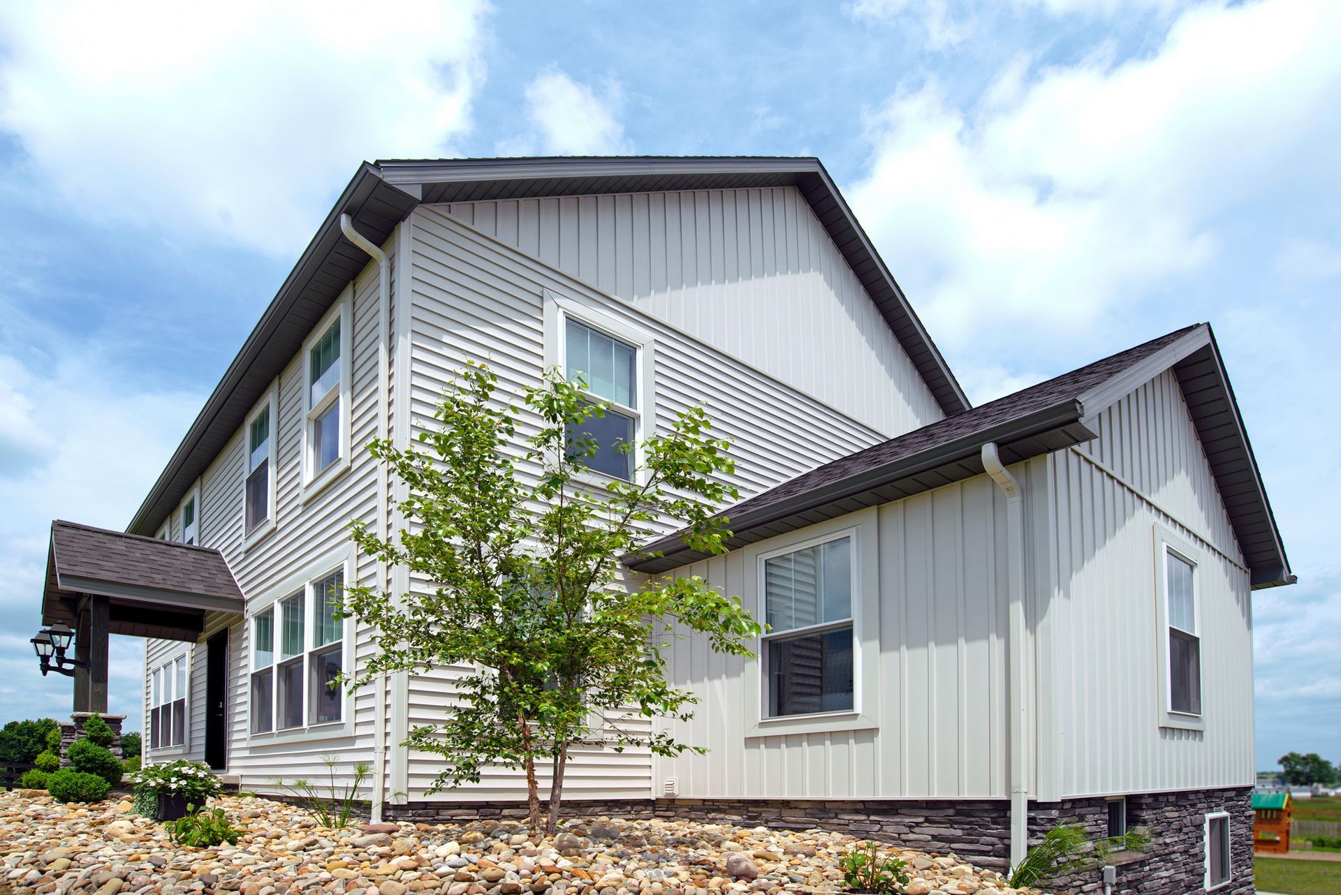 Two-story light-colored house with dark roof and trim, several windows, and small landscaping.