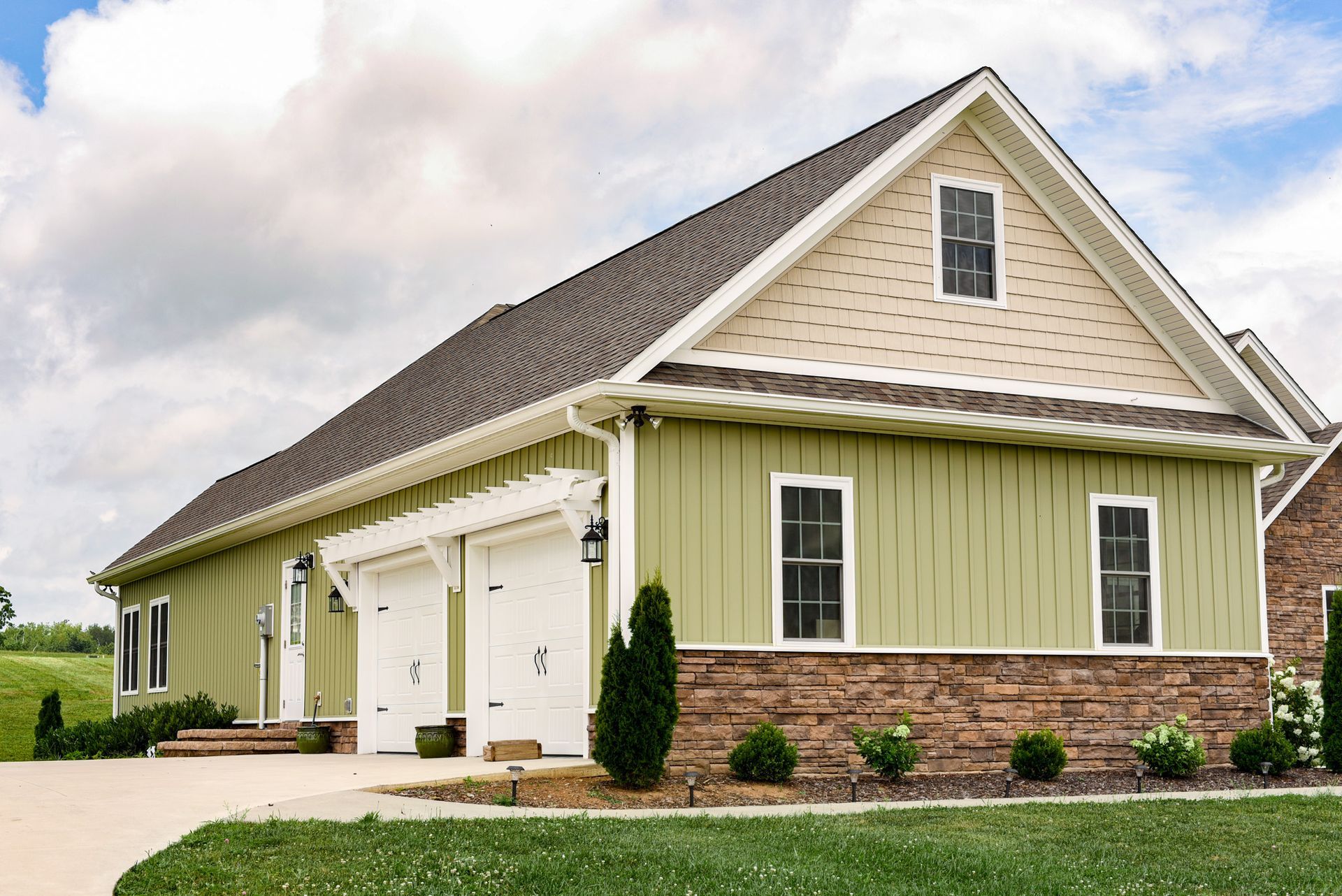 Green and stone two-car garage with white doors, tan gable, and small windows under a cloudy sky.