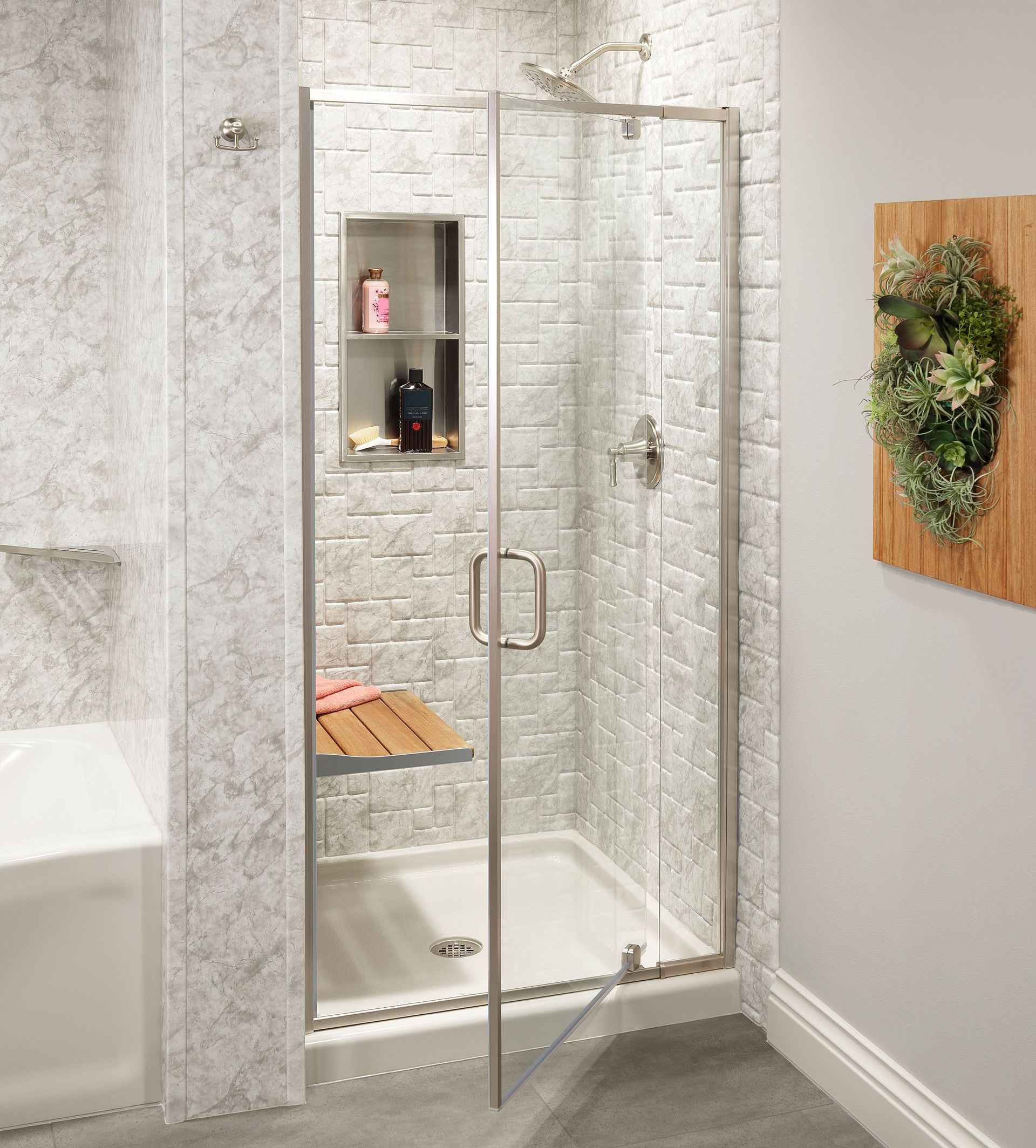 Glass shower enclosure with silver frame, built-in shelf, and wood bench. White brick-like tiled walls.