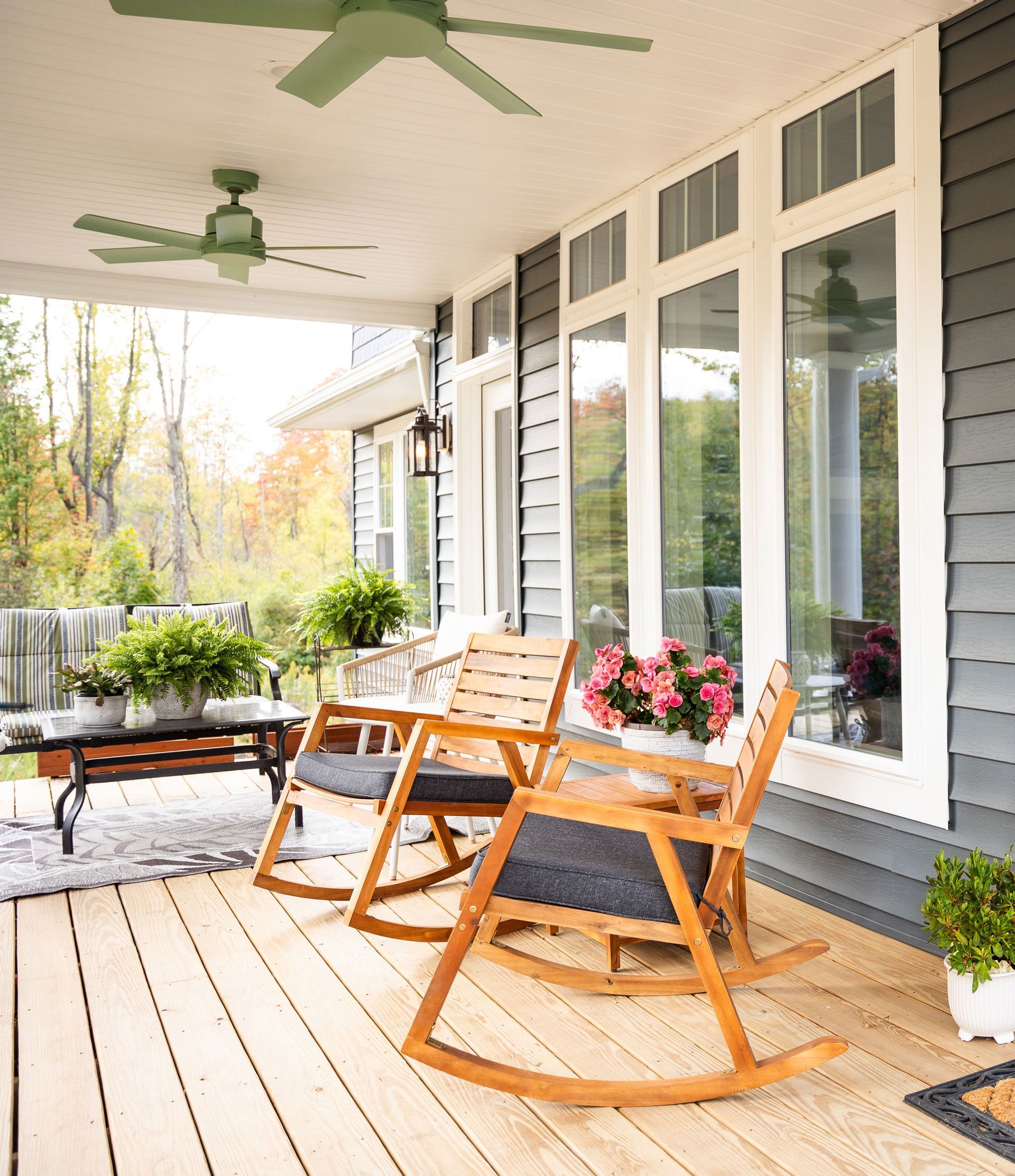 Wooden rocking chairs on a covered porch with plants and ceiling fans, overlooking a garden.