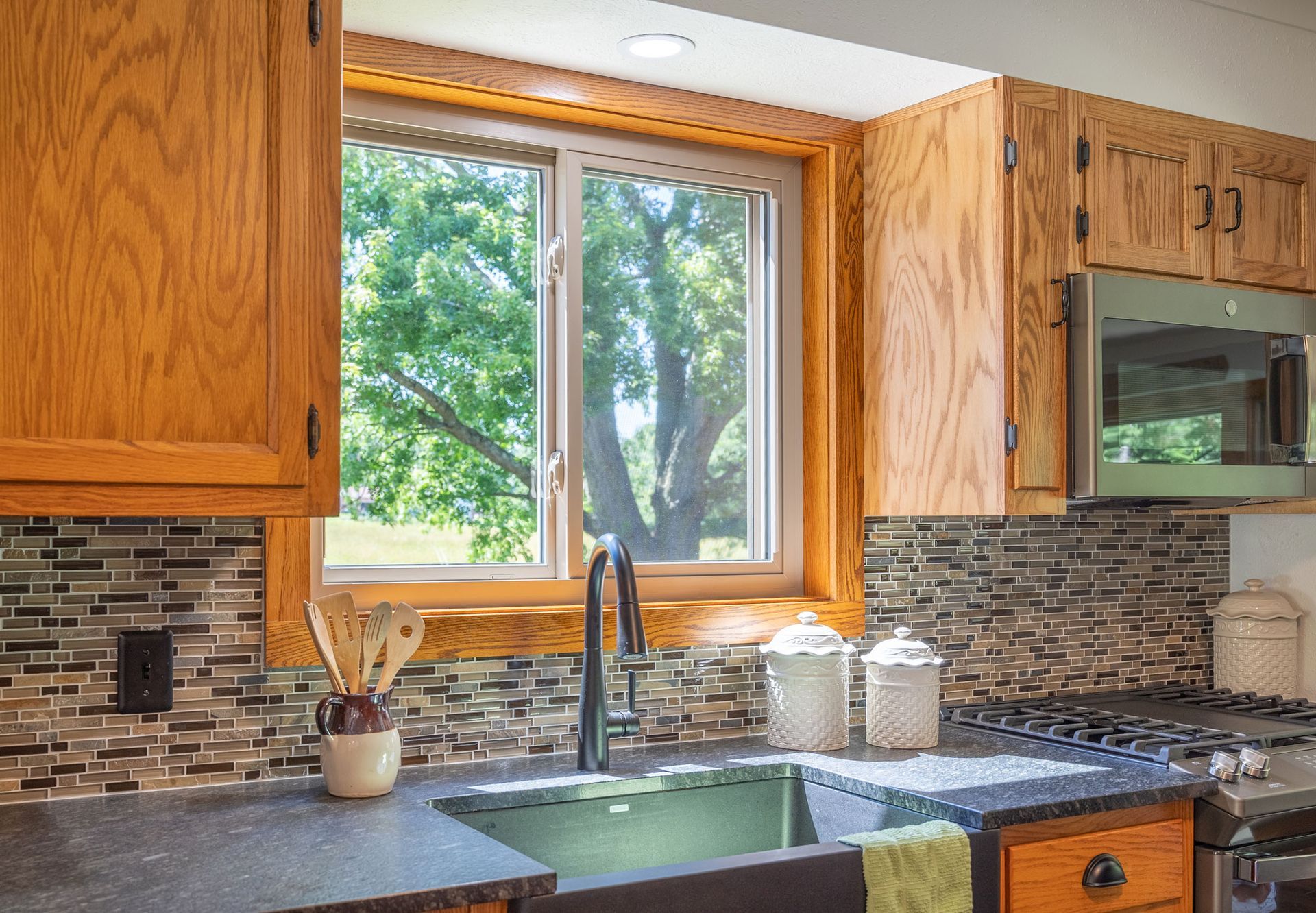 Kitchen with oak cabinets, a window with a view of trees, dark countertops, and a black sink.