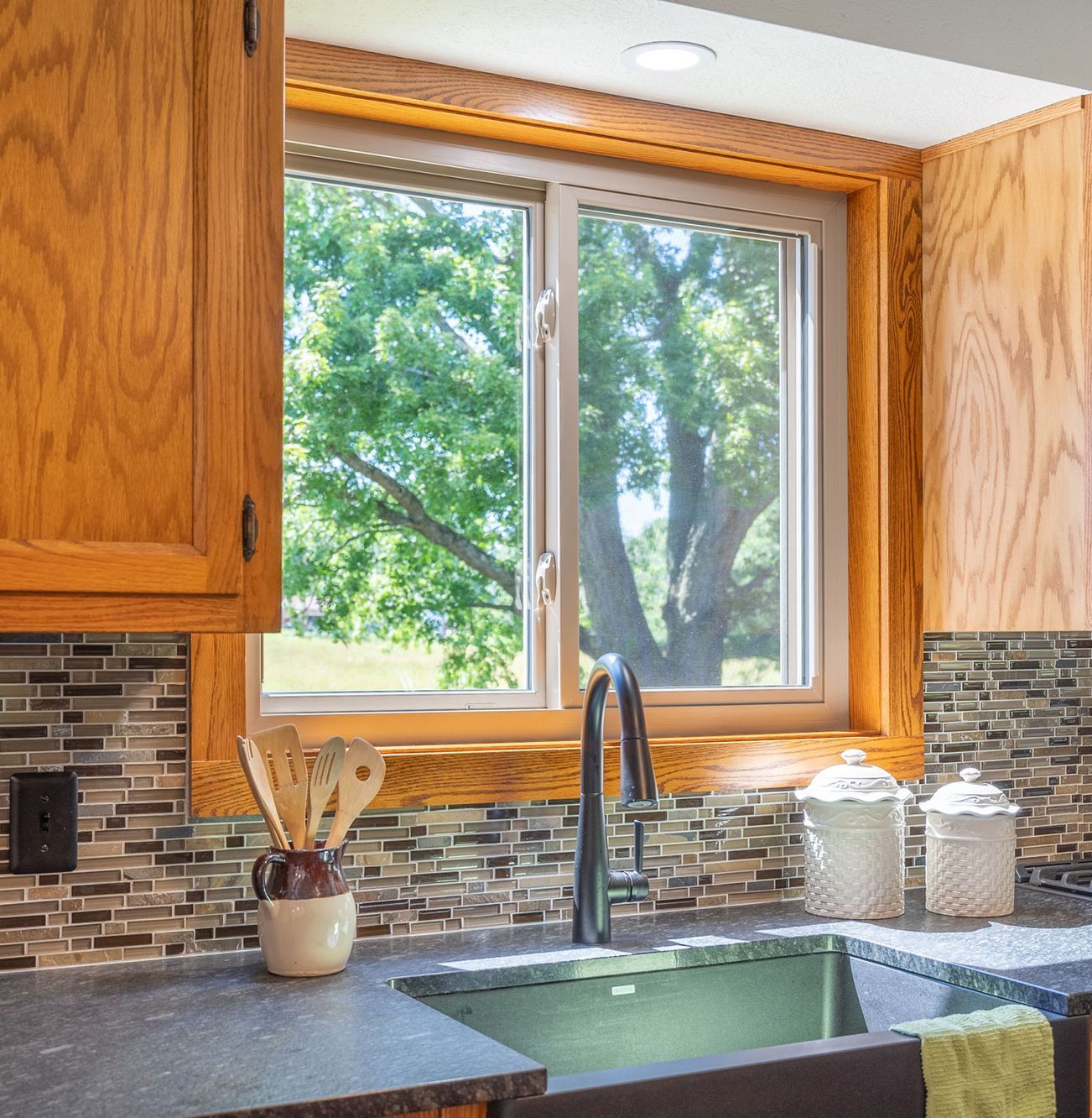 Kitchen with a window overlooking a tree, sink, wood cabinets, and mosaic tile backsplash.