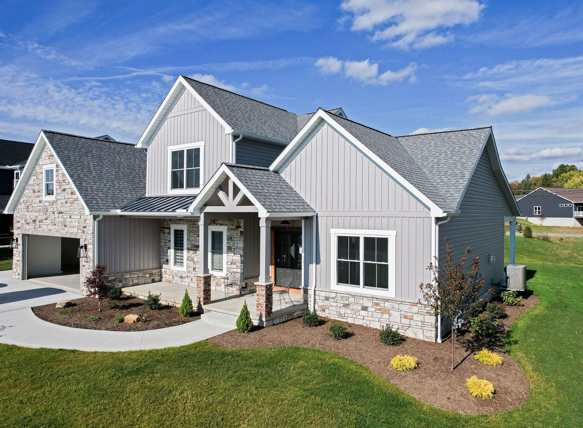 Two-story house with gray siding, stone accents, and a gray roof, set on a green lawn under a blue sky.