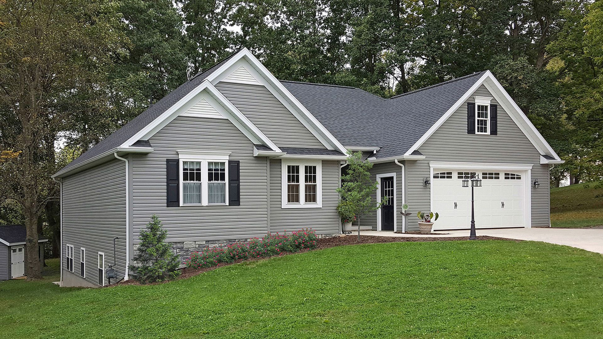 Gray house with white trim, black shutters, and a garage on a green hill.