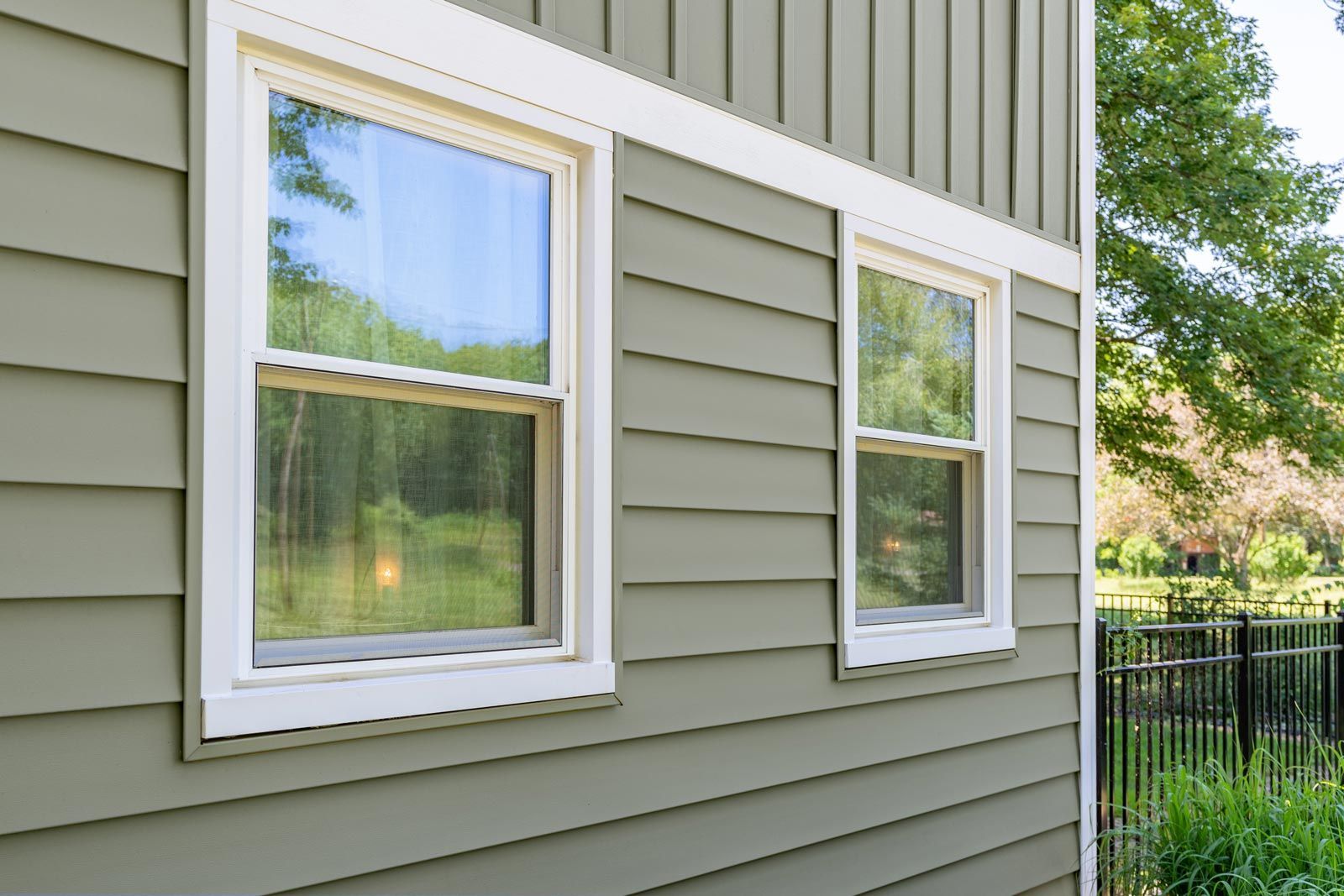 Two white-framed windows on a green-sided house, sunlight reflecting off the glass.