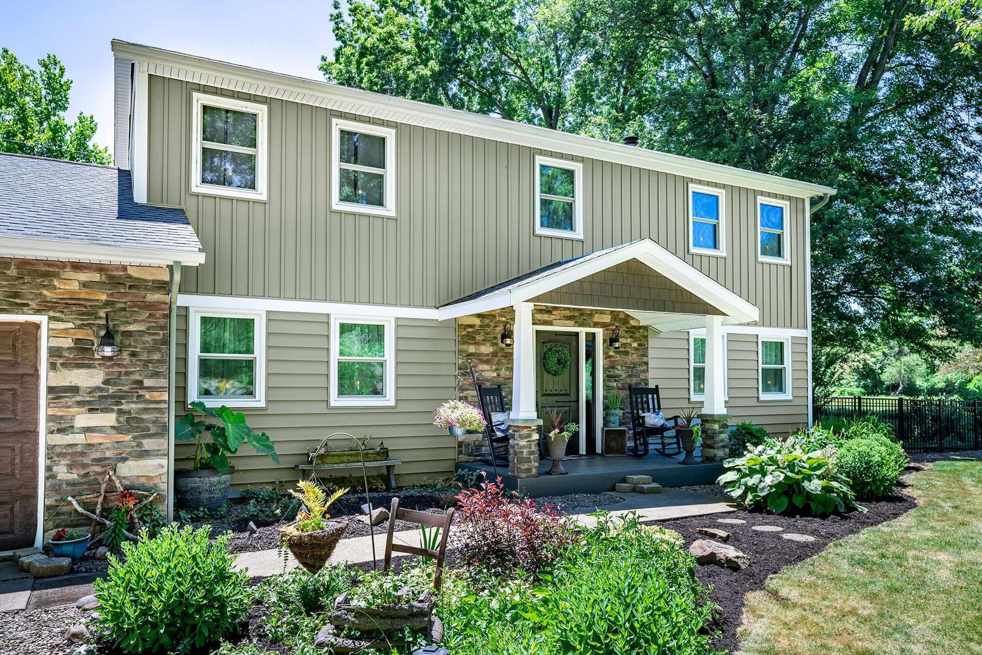 Two-story house with tan vertical siding, stone accents, and a covered porch. Lush landscaping in front.
