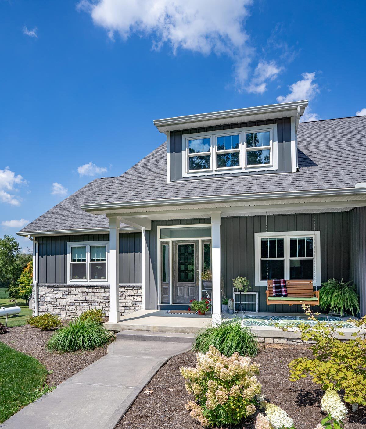 Gray-sided house with stone accents, blue sky, concrete path, and porch swing.