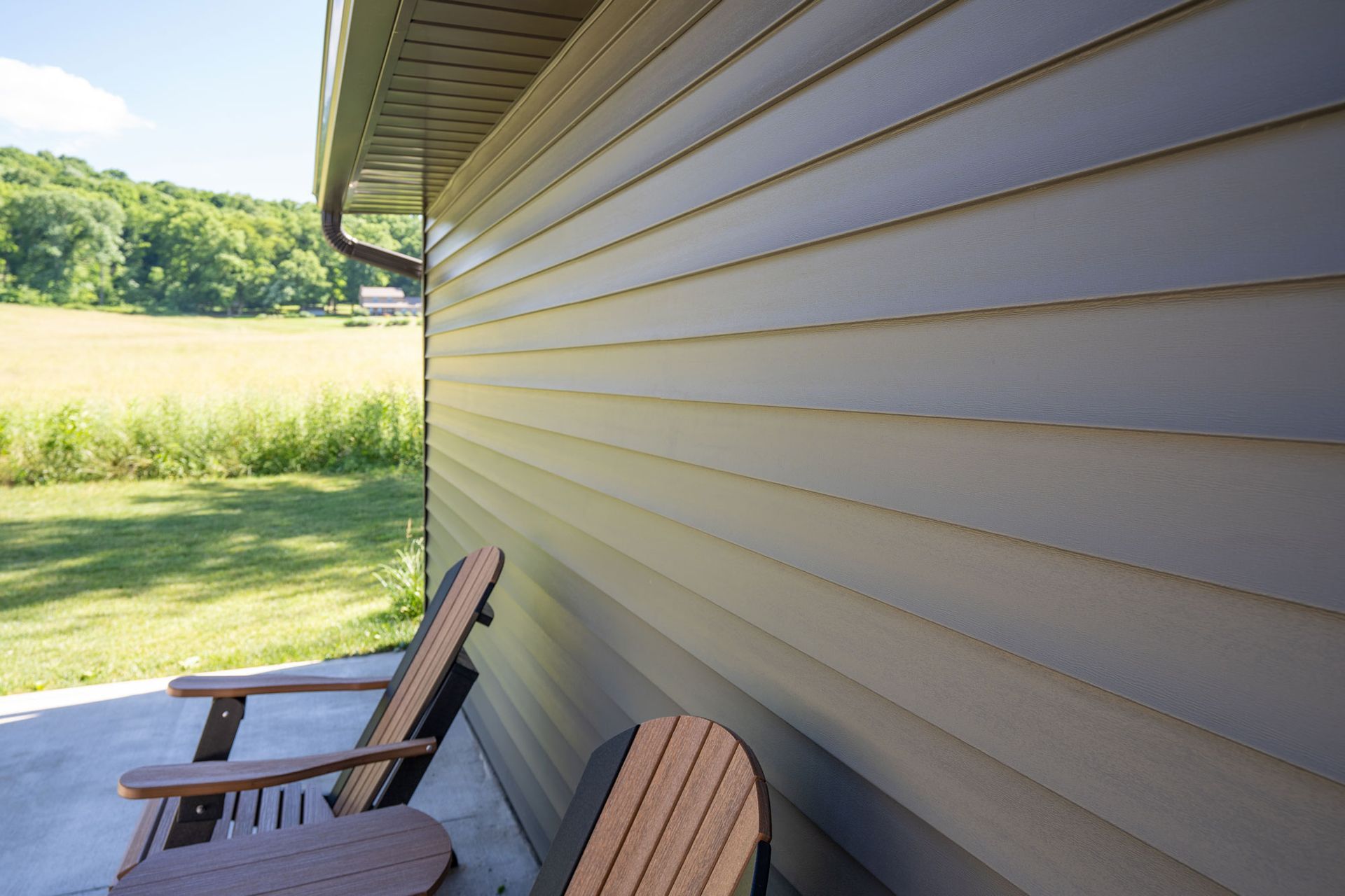Two wooden chairs on a porch with tan siding on a building, field and trees in the background.