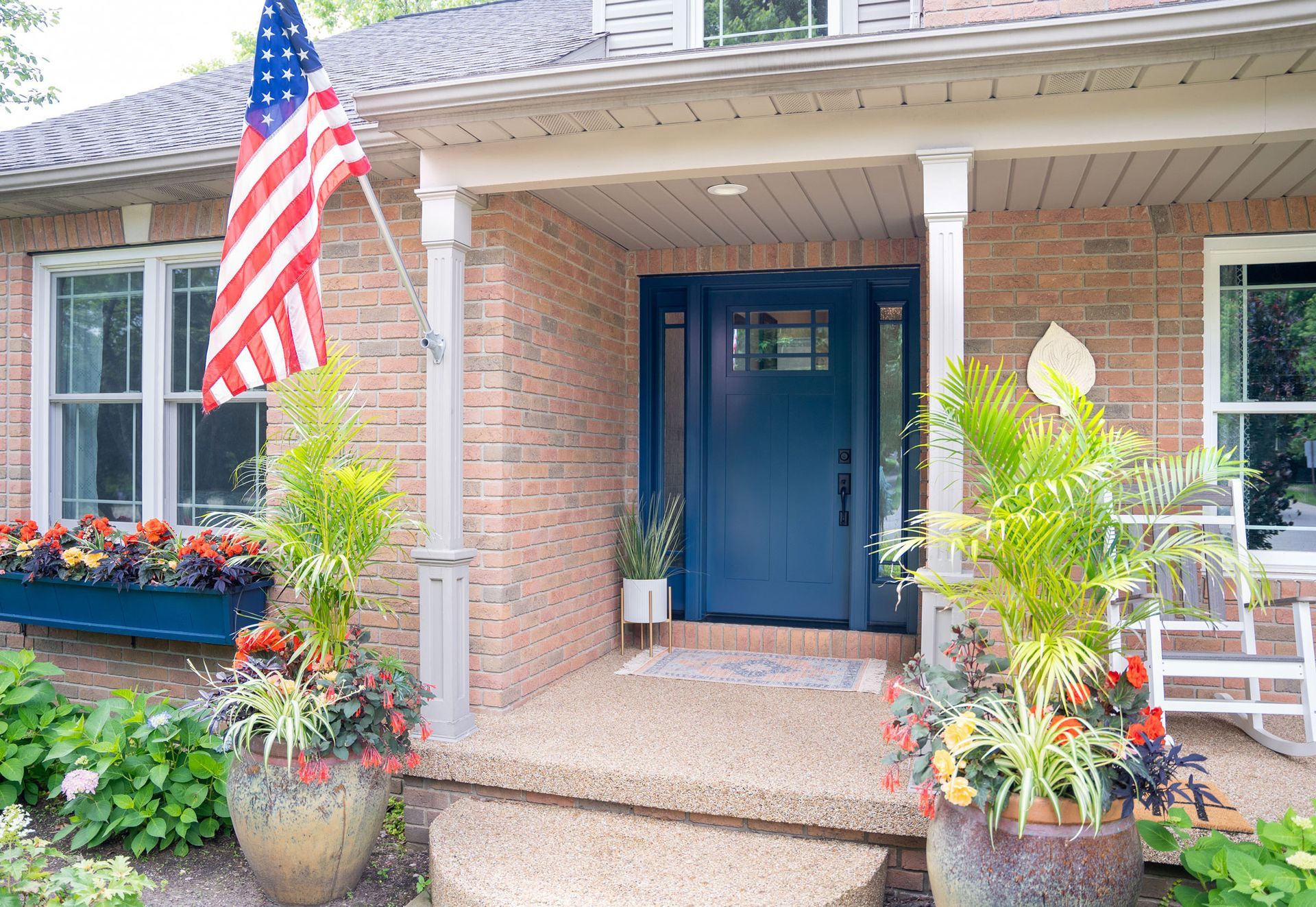 Brick home entrance with navy blue door, American flag, and potted plants.