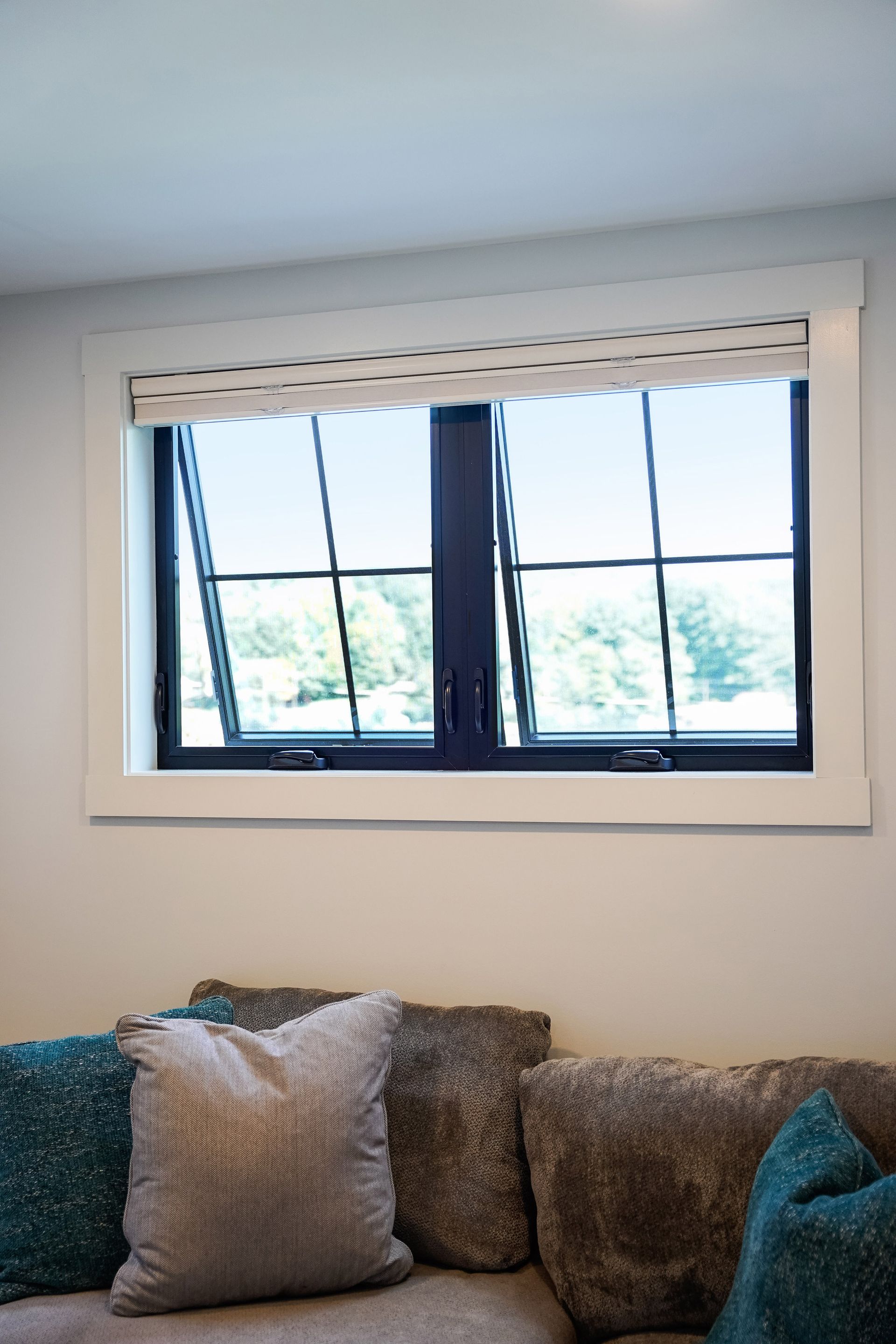 Window with black frames, white trim, and a patterned shade, above sofa with pillows.