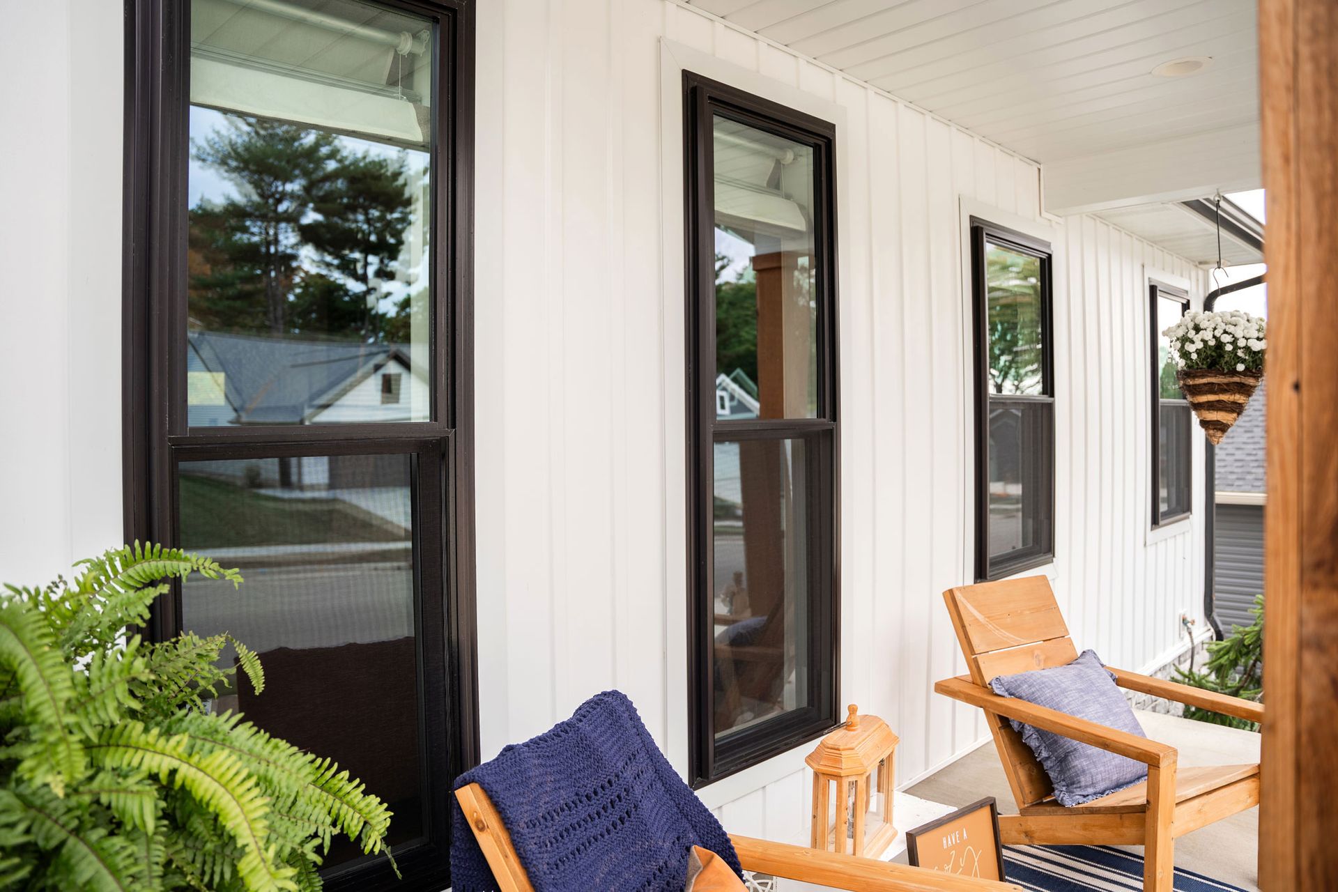 White porch with black-framed windows, wooden chairs, and ferns.