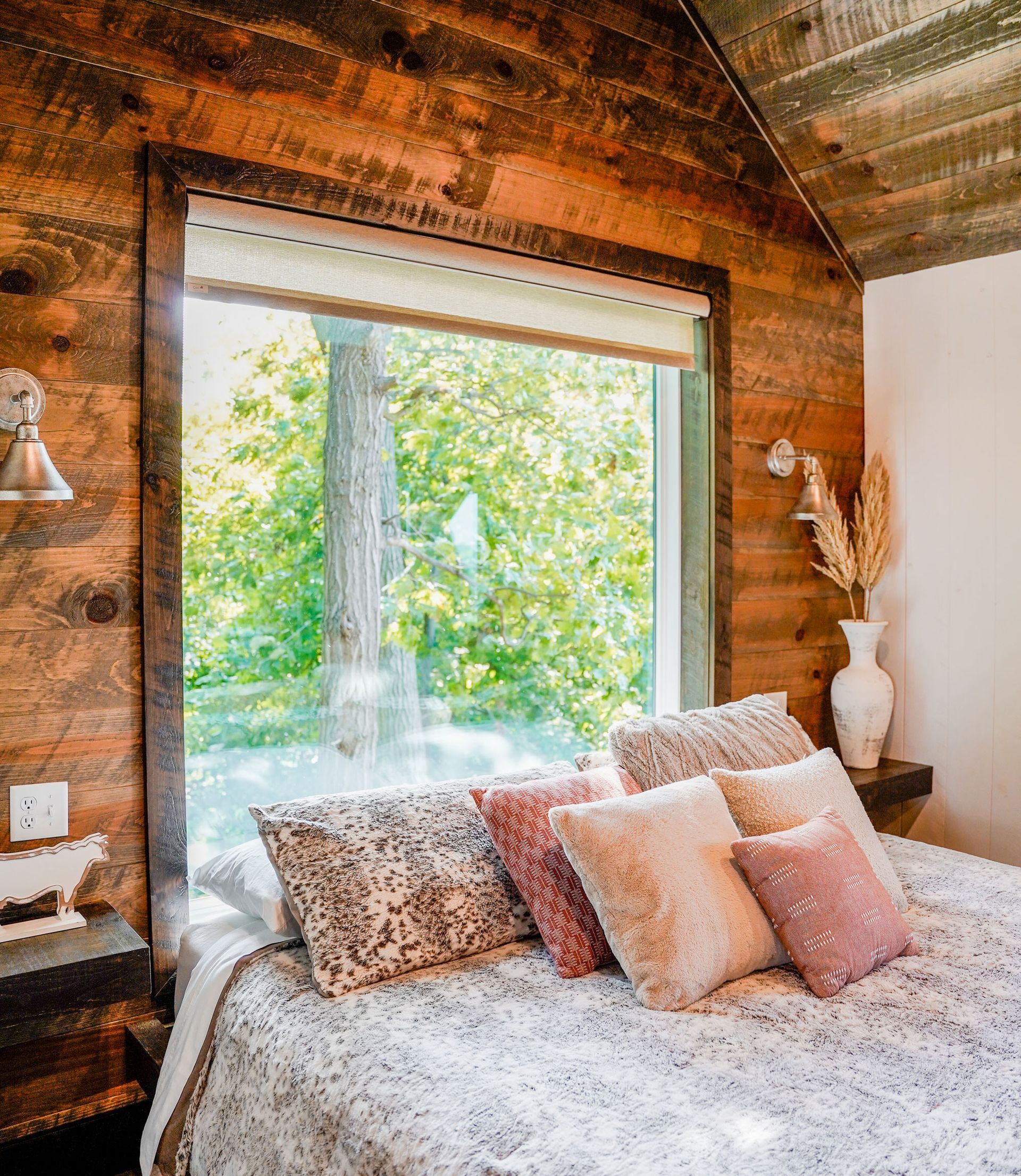 Bedroom with wooden walls, large window overlooking trees, bed with pillows.