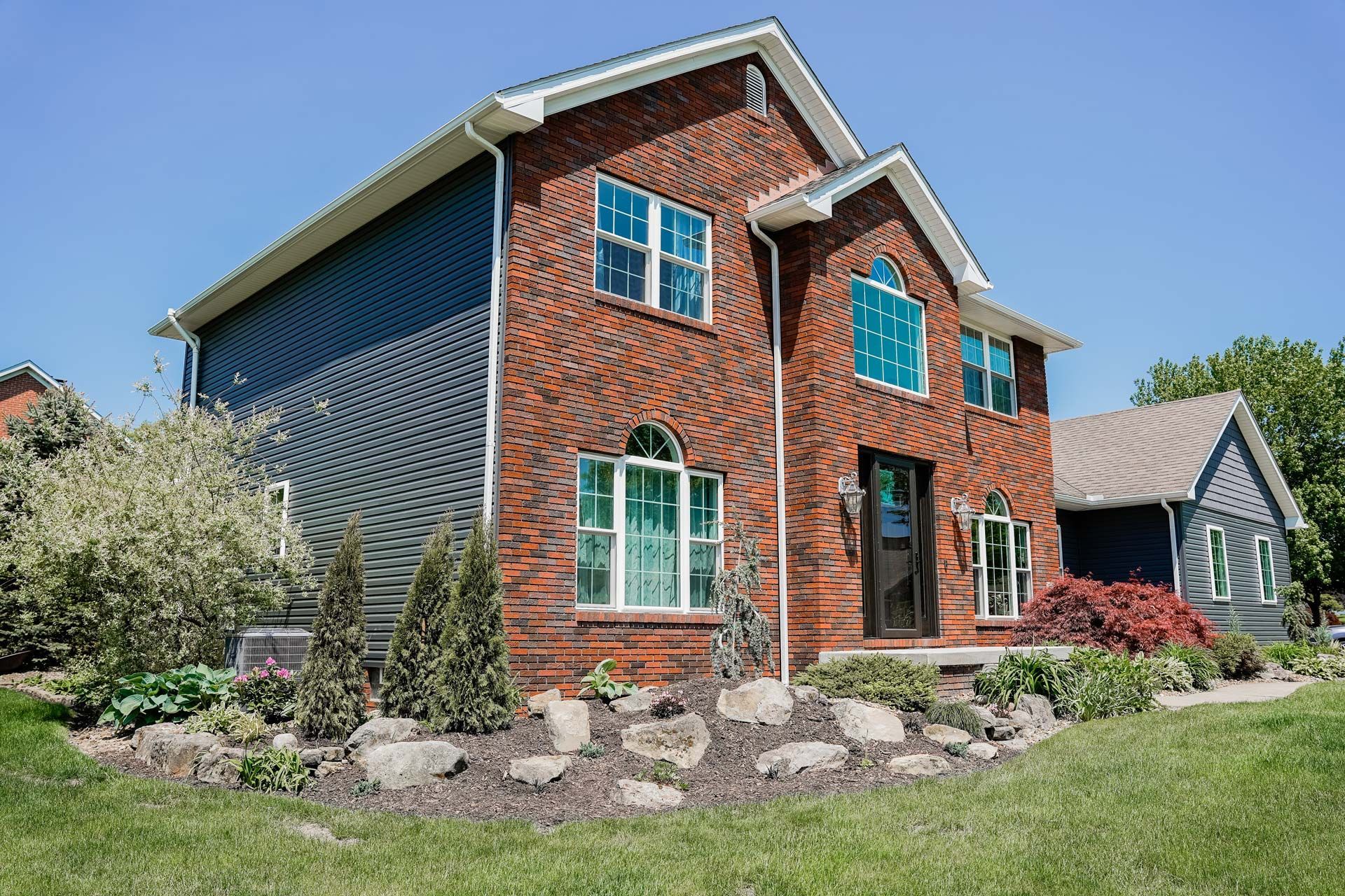 Two-story brick and blue-sided house with landscaping and a sunny sky.