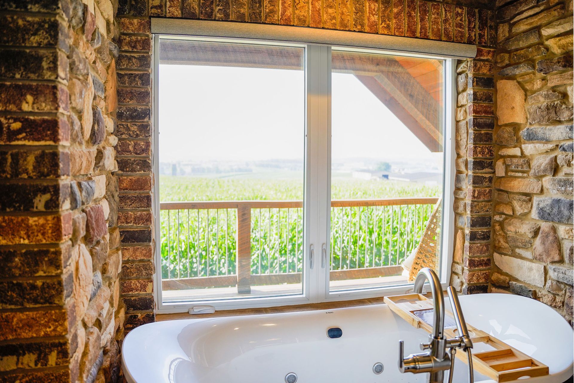 Bathroom with a white bathtub by a window overlooking a field, surrounded by brick walls.
