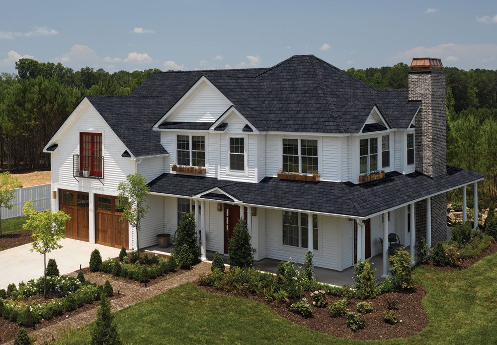 White farmhouse with black roof, wrap-around porch, and stone chimney.