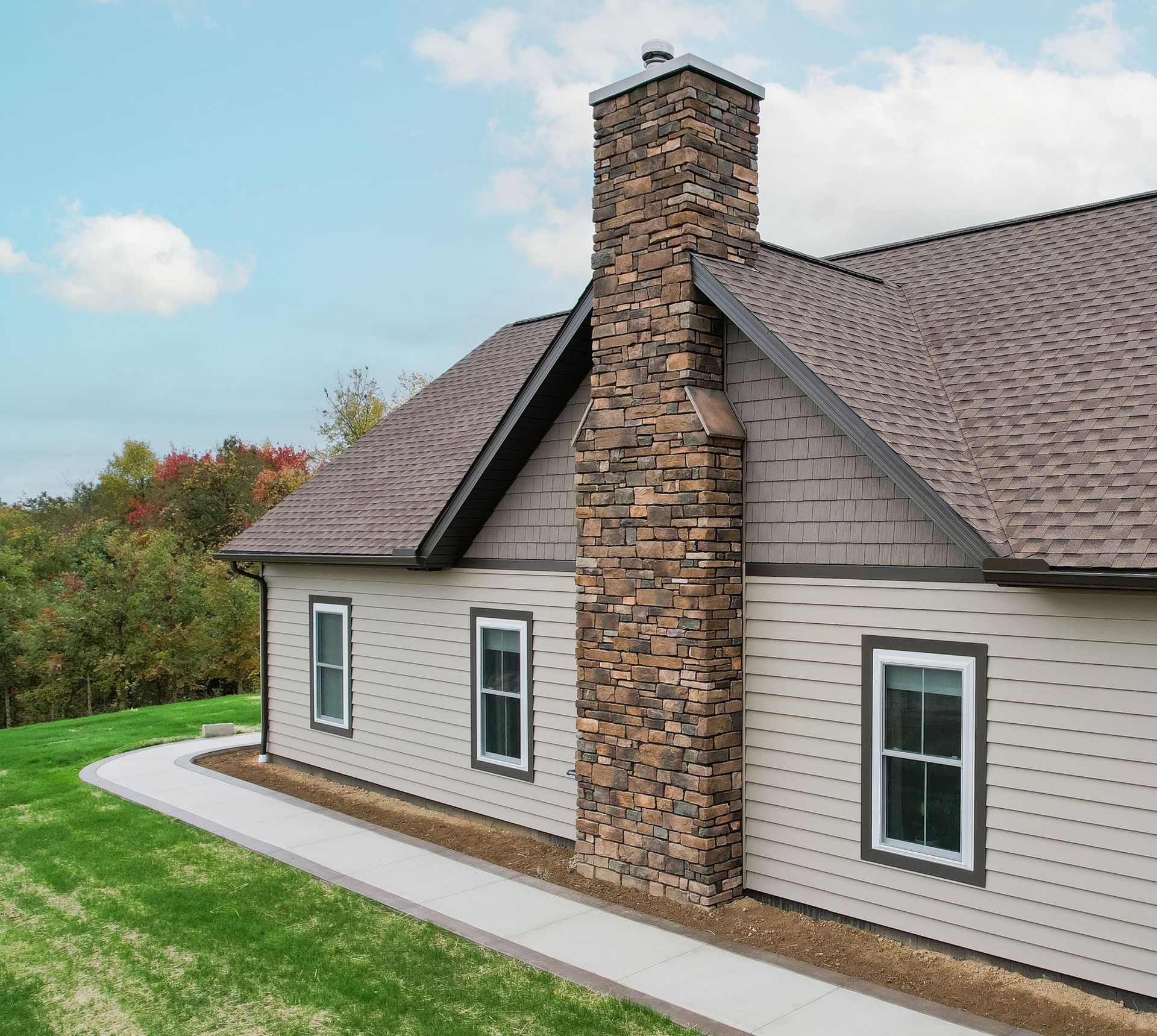 Tan house with stone chimney, brown roof, and white-framed windows on a sunny day.