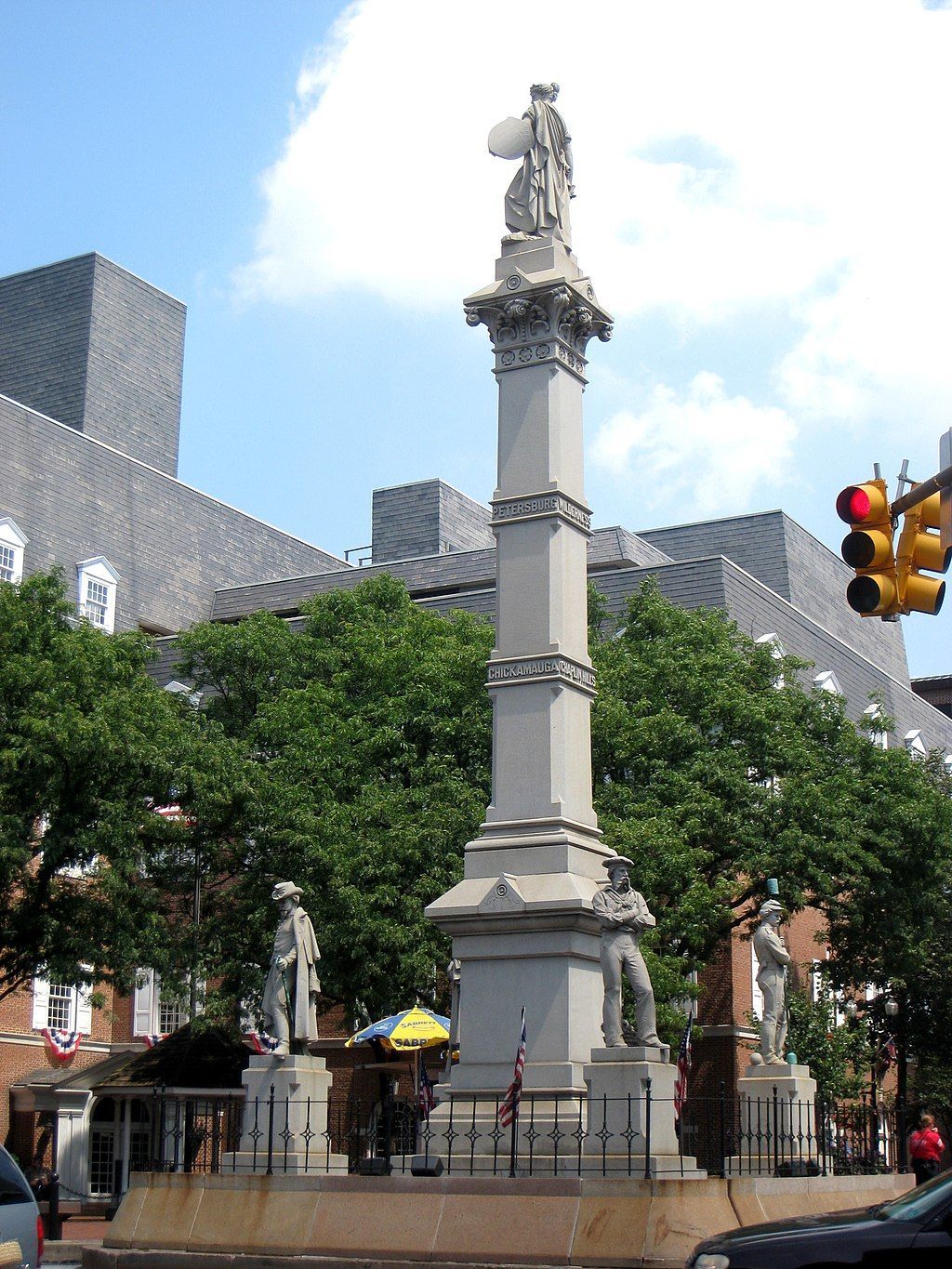 Monument with statues in public square, trees and building in the background. Red traffic light visible.