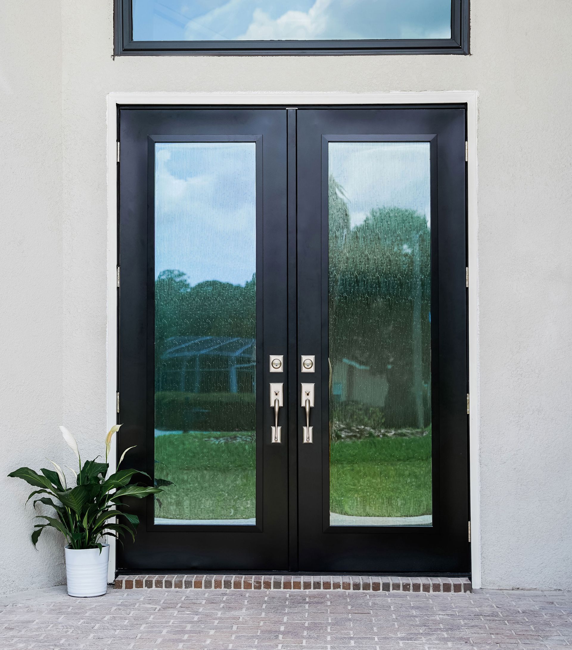 Black double doors with glass panels and silver hardware, set in a light-colored stucco wall with a brick threshold.
