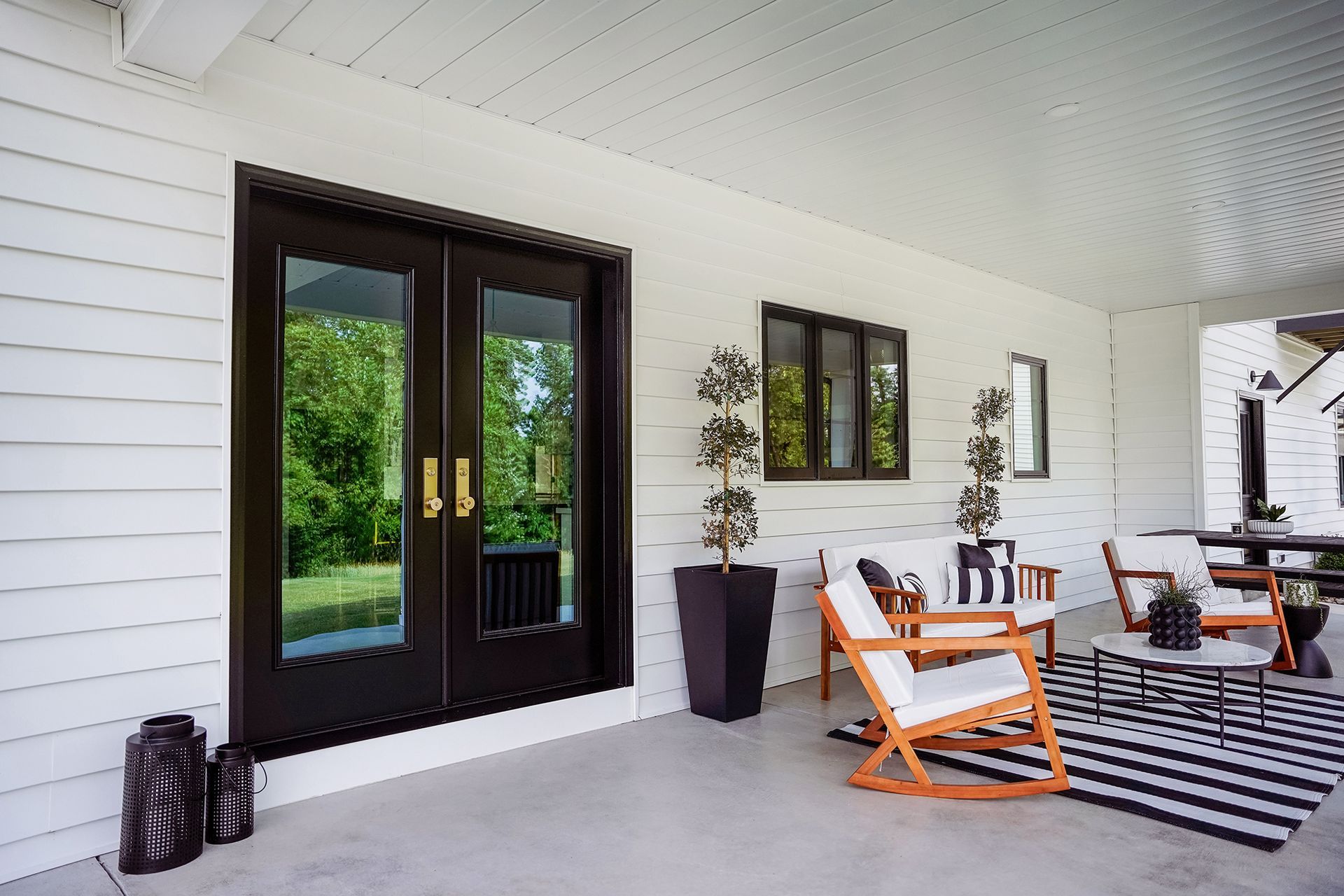 Covered porch with black-framed French doors, outdoor seating, black and white rug, and potted plants.