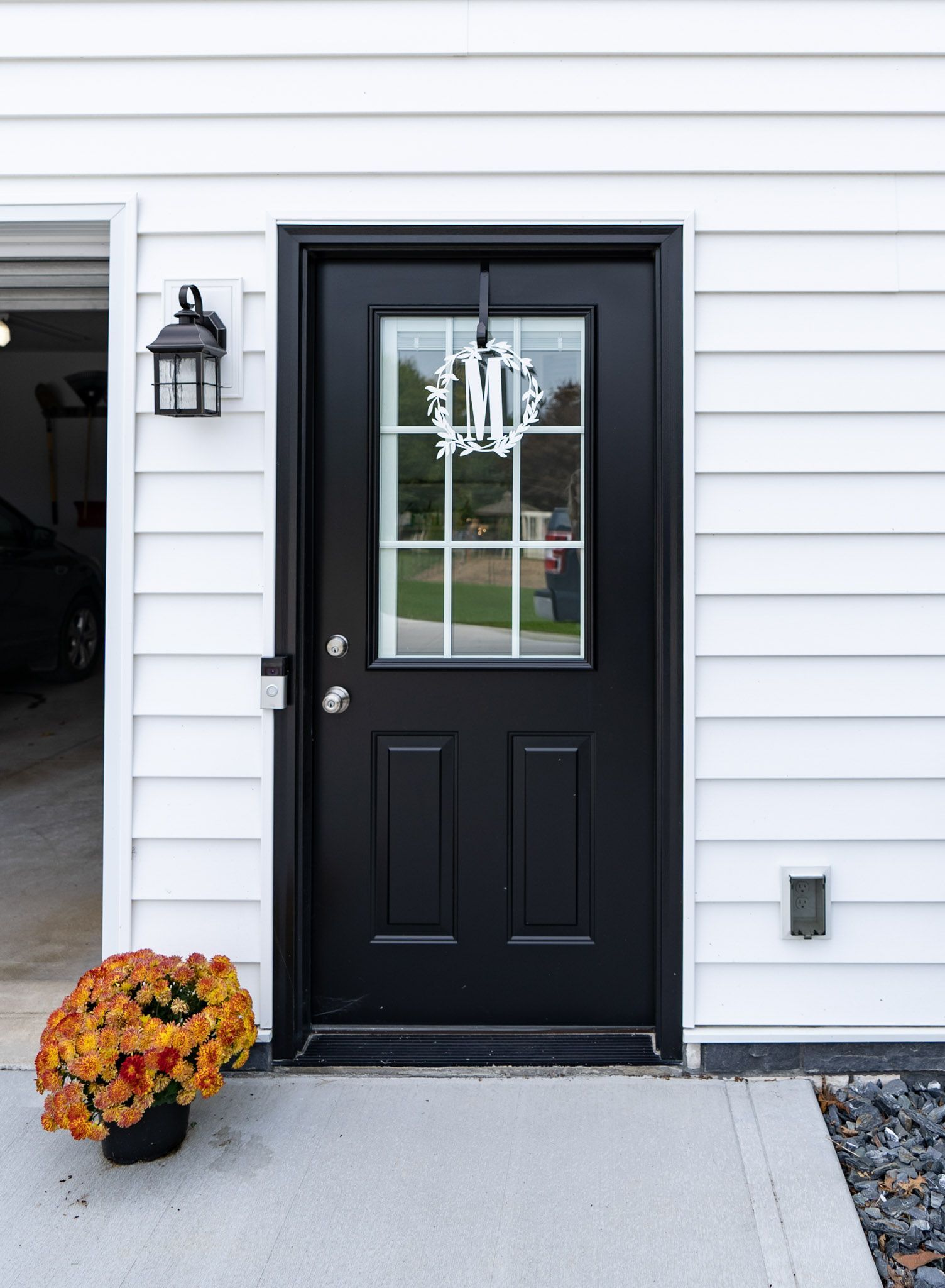 Black door with glass panel and wreath, white siding, lantern, and orange flowers.
