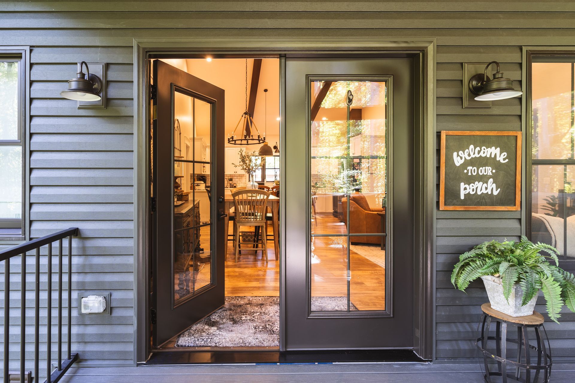 Open brown glass doors reveal a warmly lit interior. Gray siding, outdoor lights, and a welcome sign decorate the exterior.