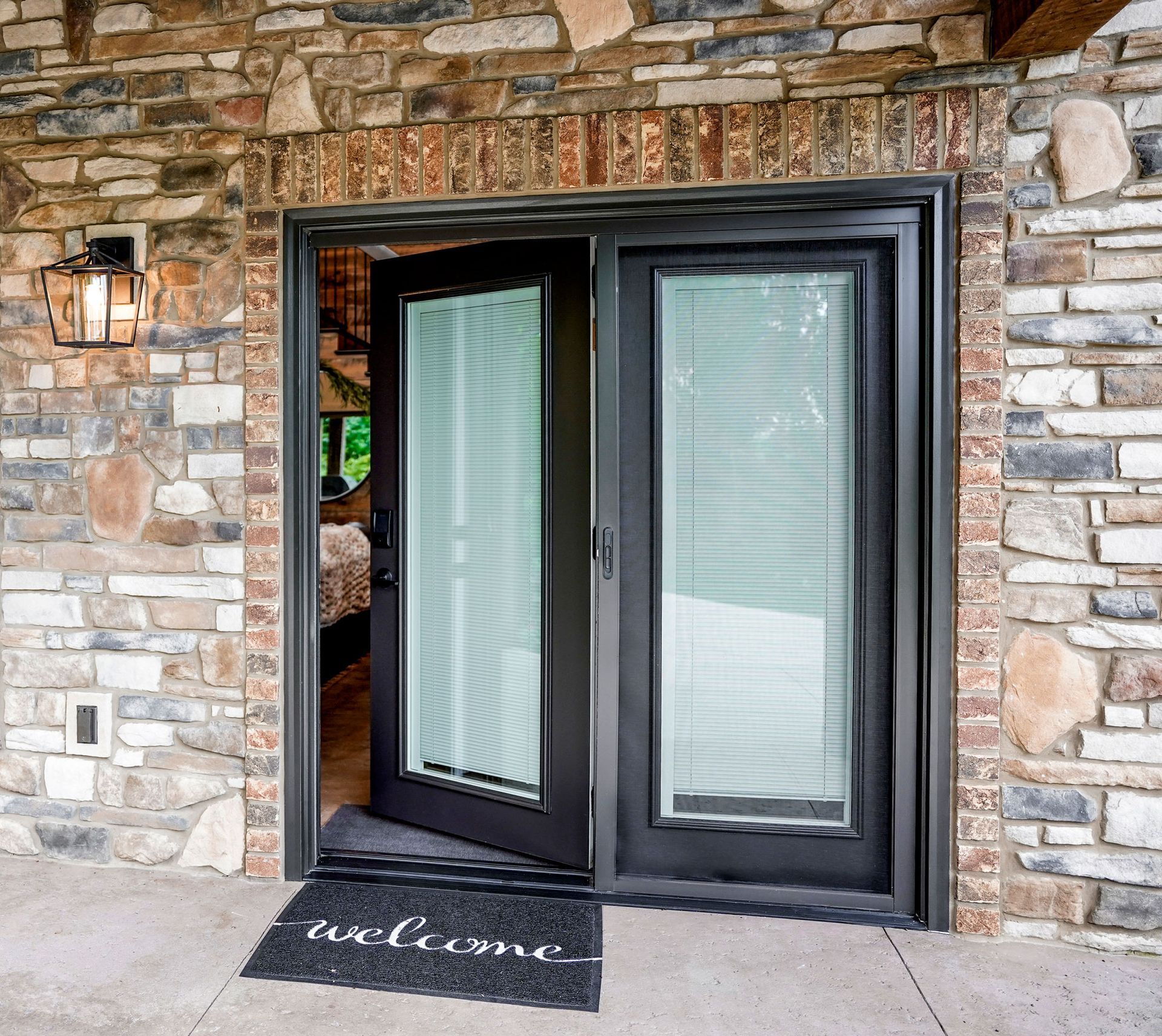 Black double doors with blinds, set in a stone wall, open to a room. A welcome mat is on the ground.