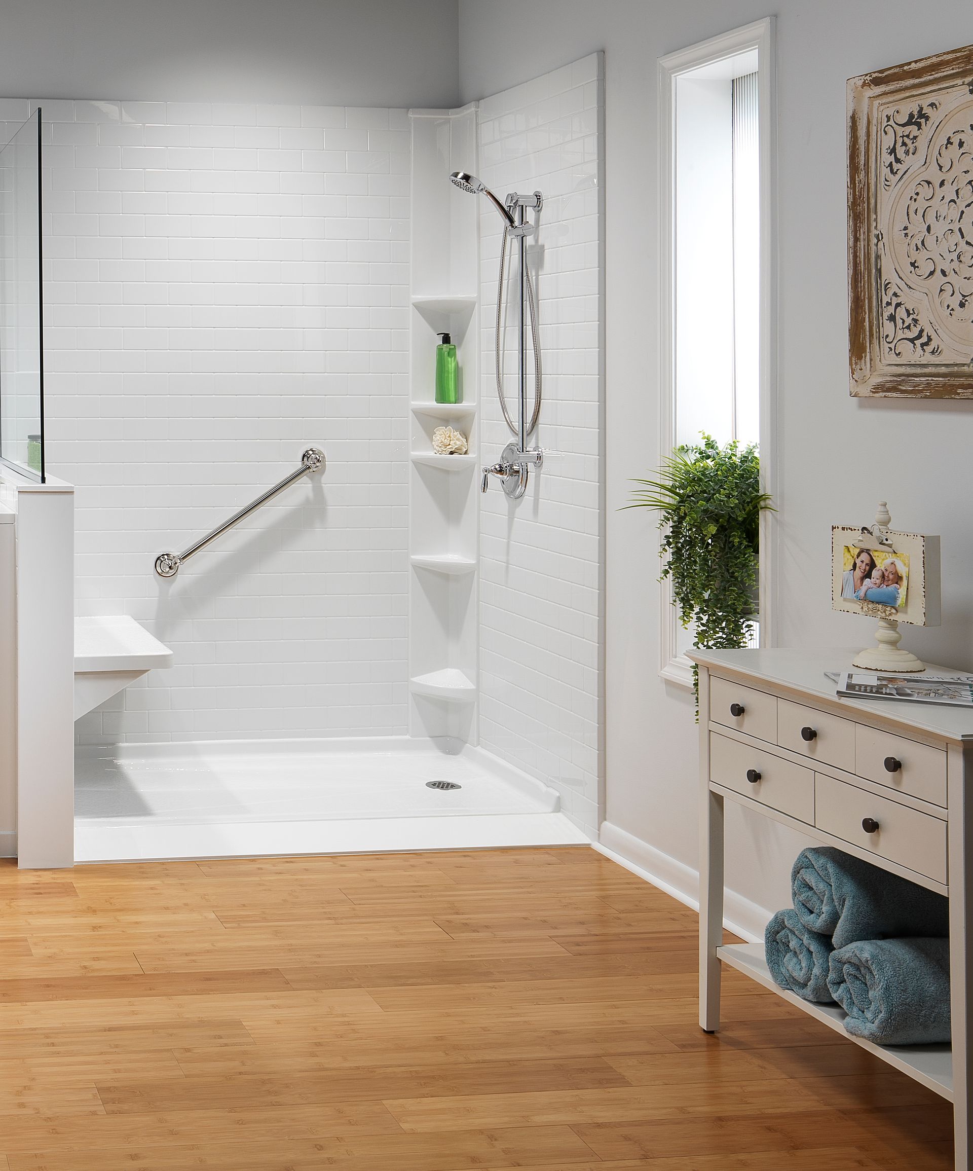 White accessible shower with built-in seat, grab bar, and shelving. Cork floor, window, and small cabinet.