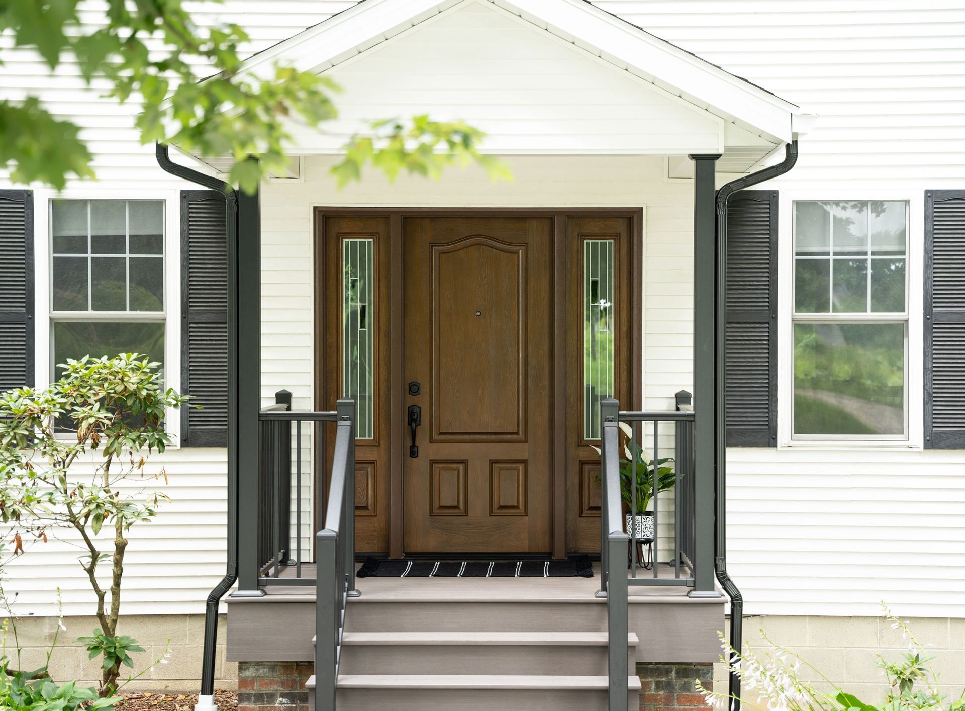Brown front door with sidelights and steps leading up to a white house with black shutters.