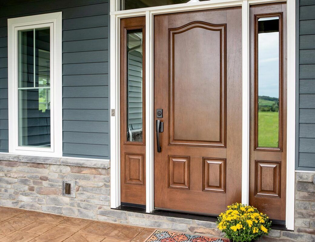 Brown wooden front door with sidelights, flower pot, and blue siding.