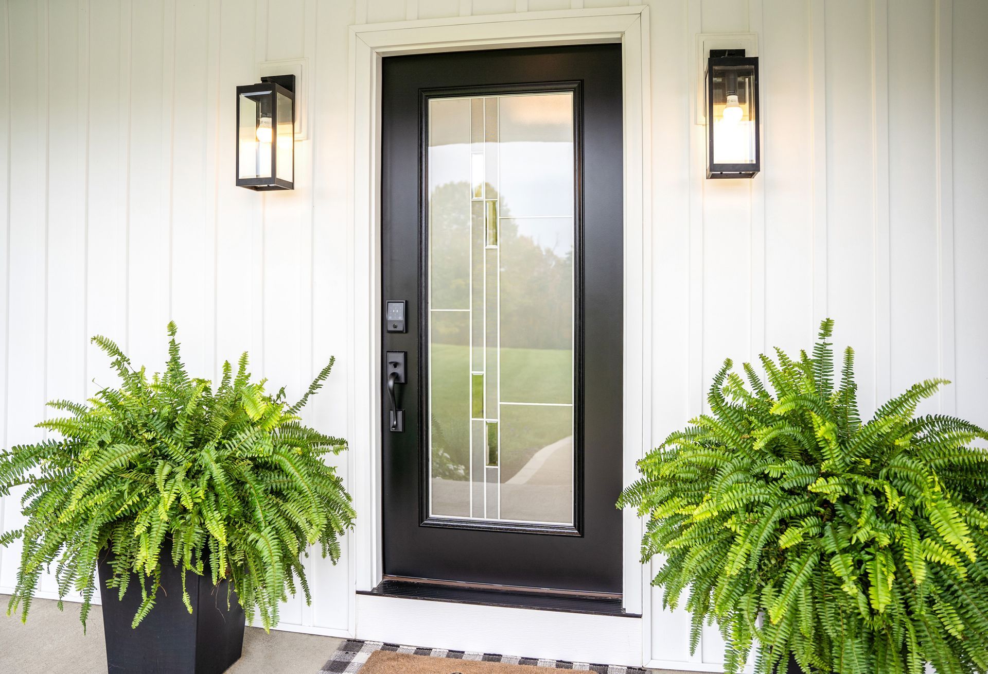 Black front door with glass panel, flanked by ferns and wall-mounted lights on a white house.