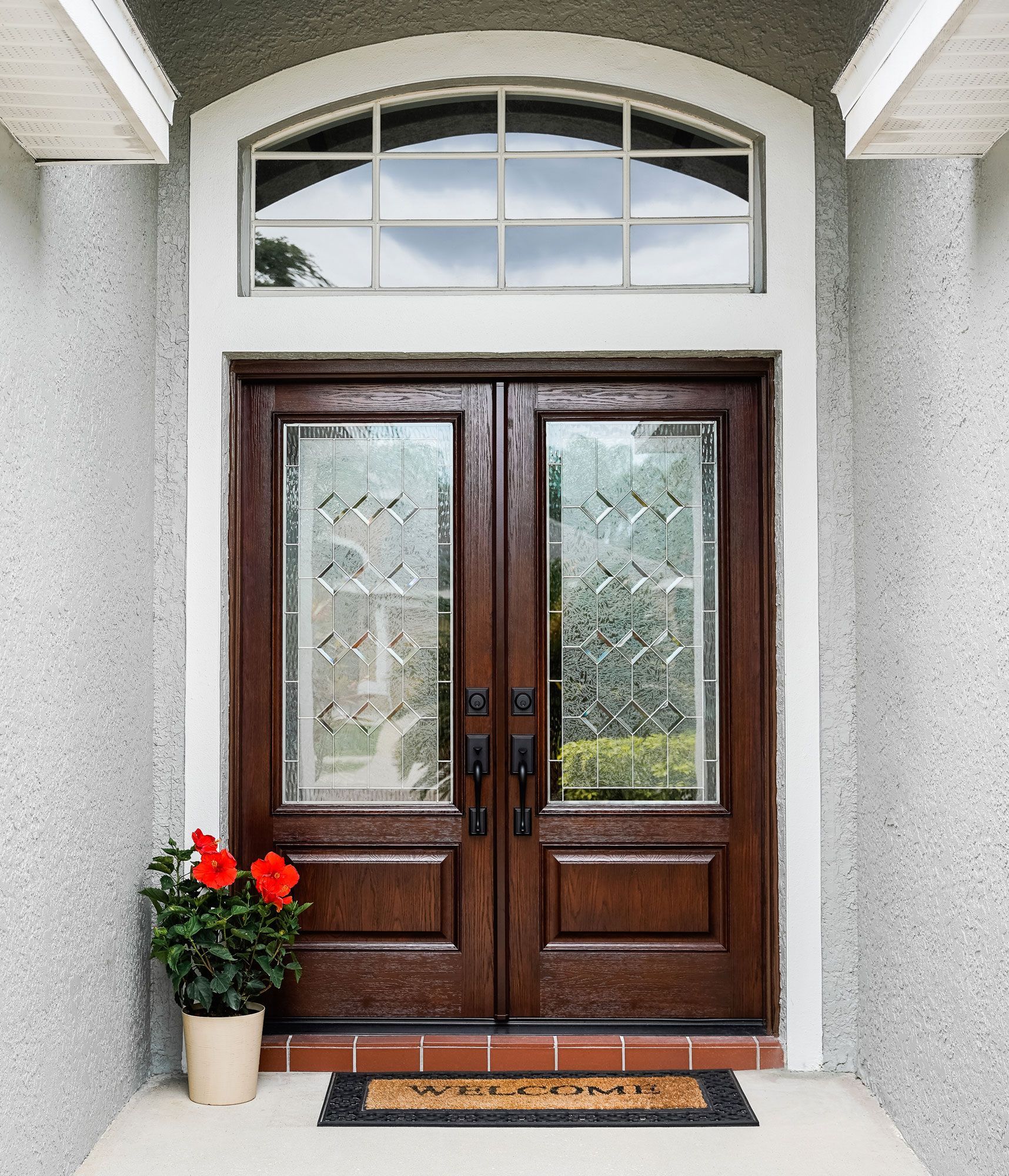 Double brown front doors with beveled glass and an arched window above. Red flowers in a pot, welcome mat.