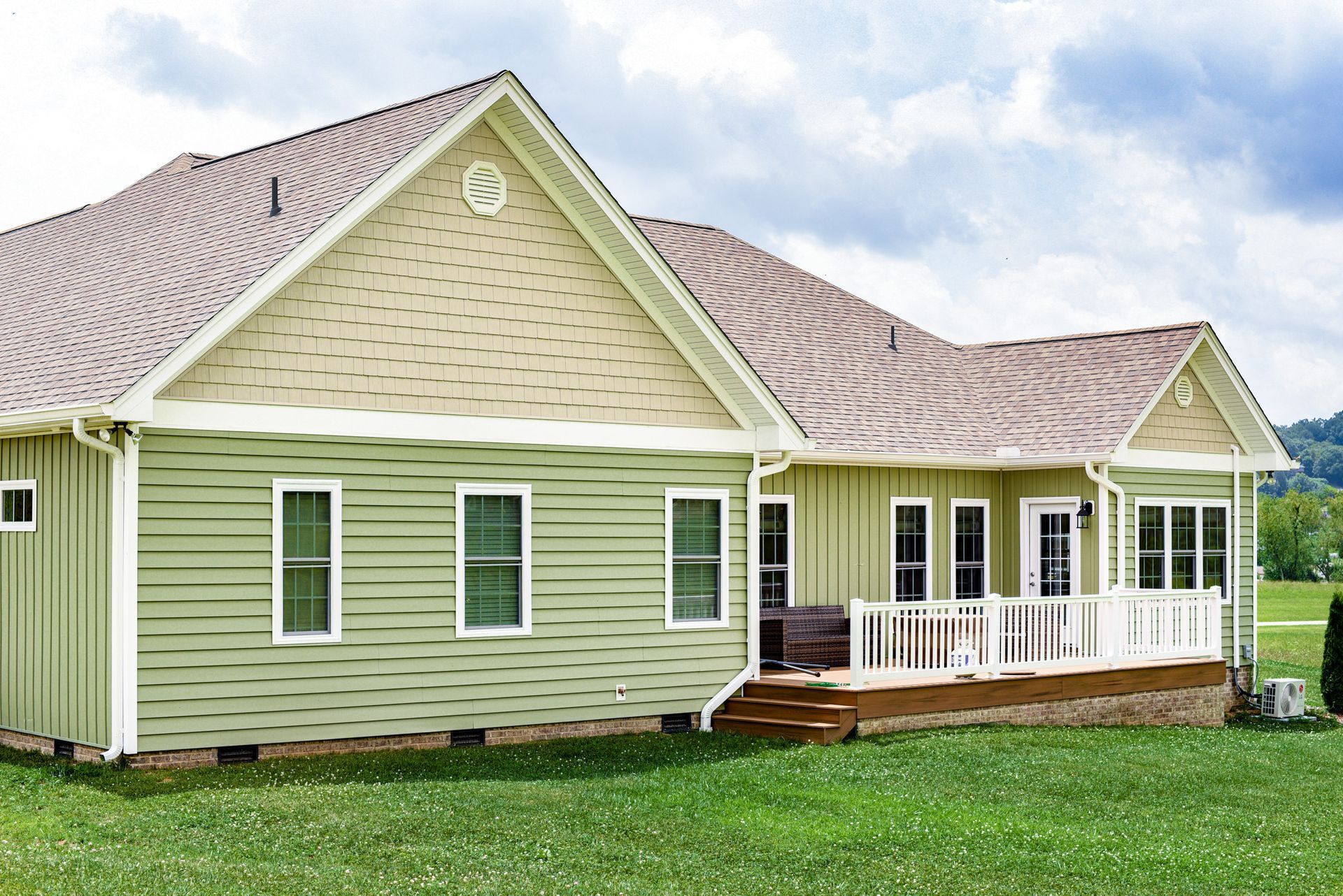 Green-sided house with a porch and brown roof on a grassy lawn under a cloudy sky.