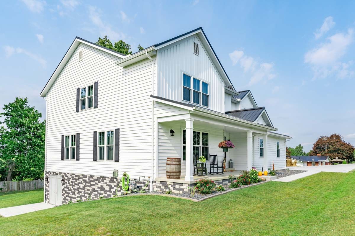 White farmhouse with black shutters and porch, on a grassy hill under a blue sky.