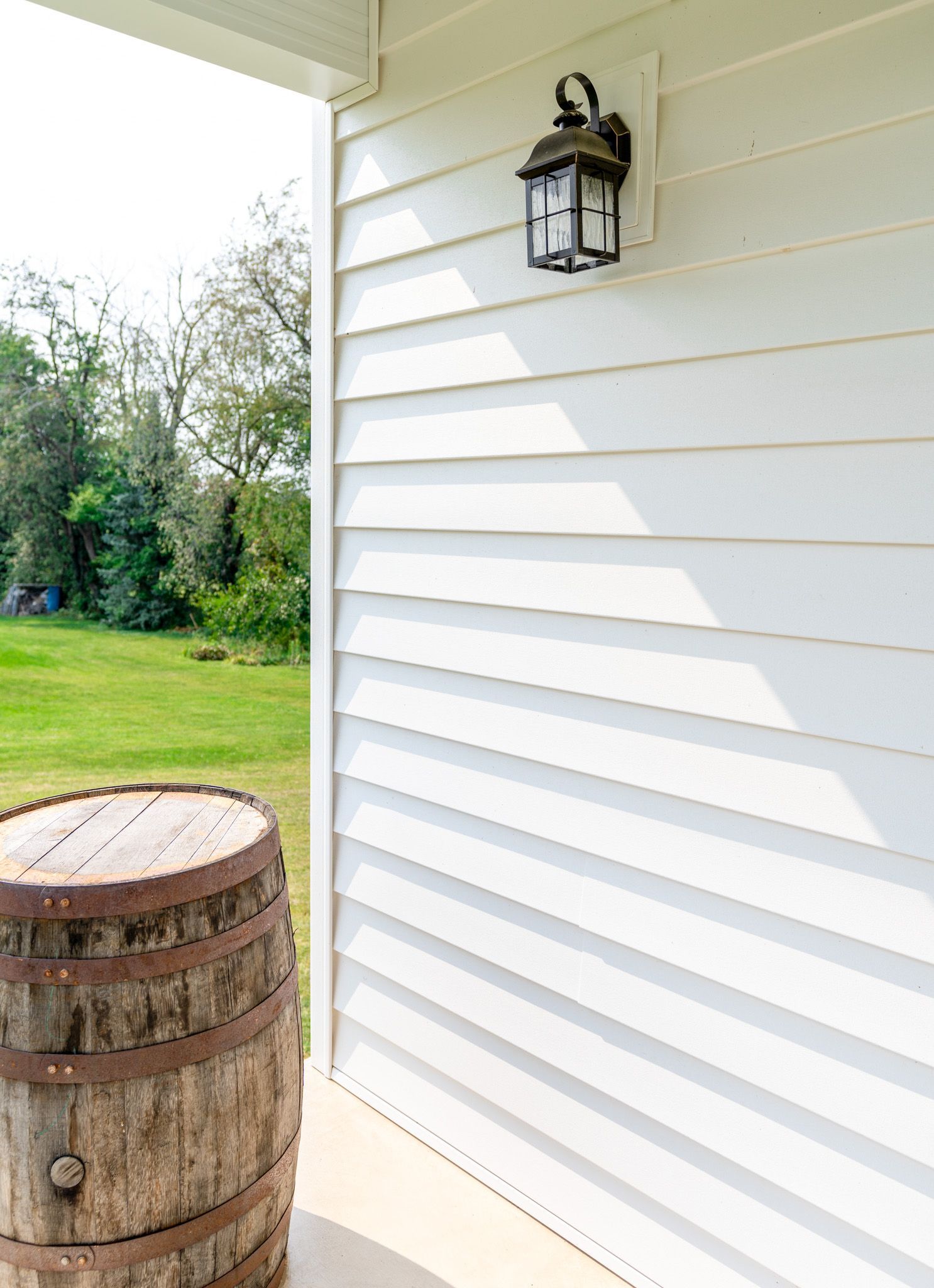 Wooden barrel on porch next to white siding with a black lantern. Green grass and trees in the background.