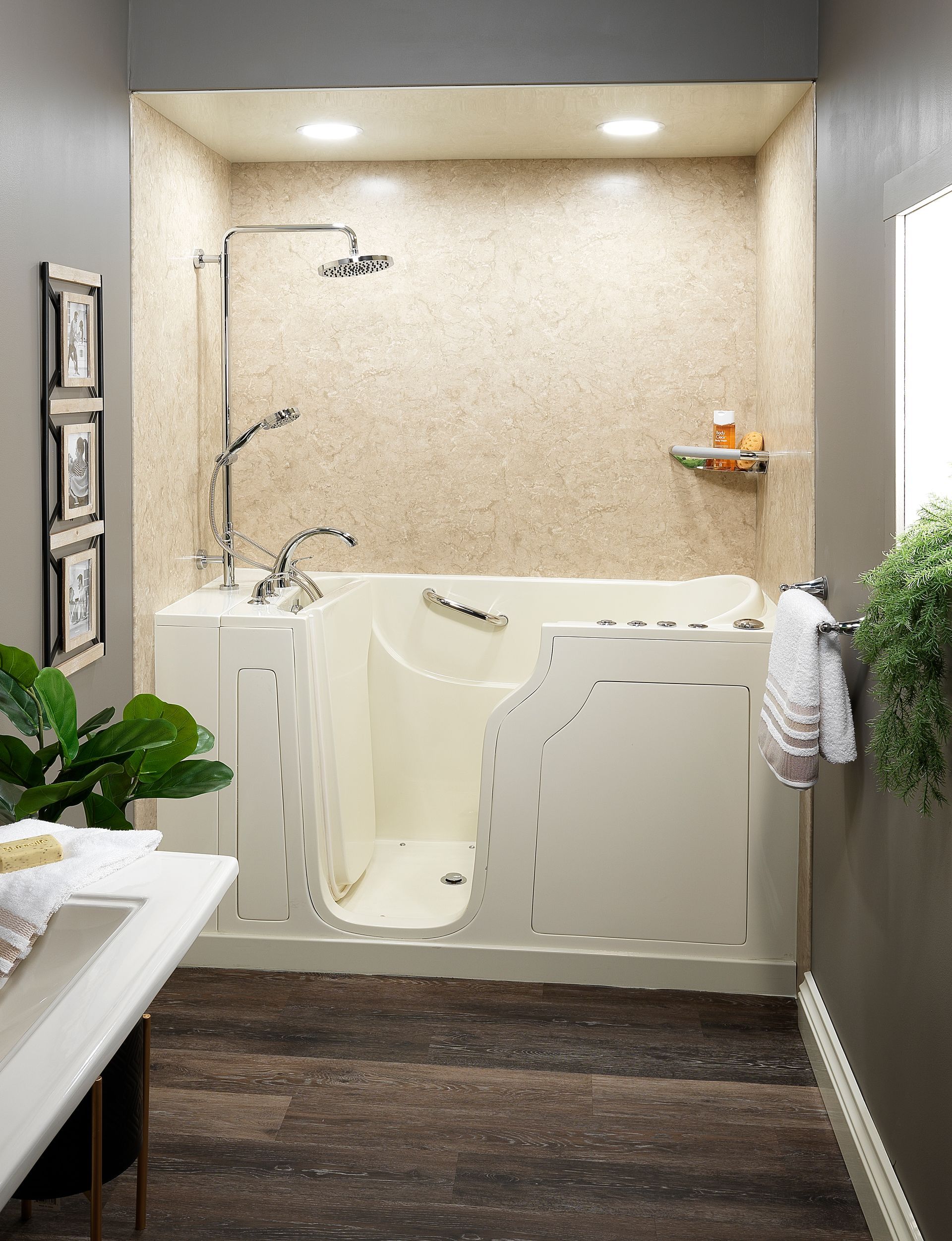 Walk-in tub in a beige alcove with showerhead and grab bar, next to a white sink and greenery.