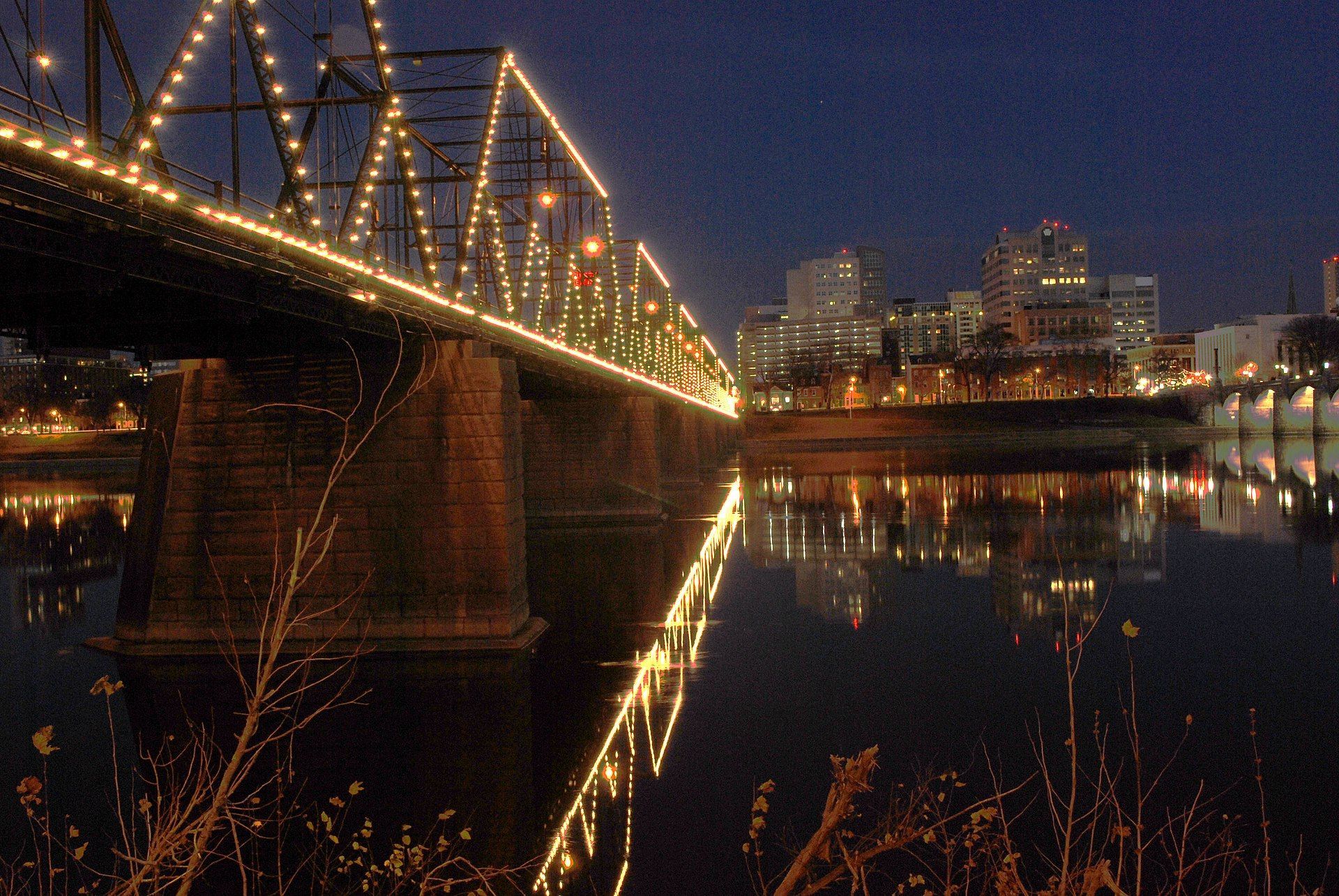 Bridge with string lights over water reflecting city lights at night.